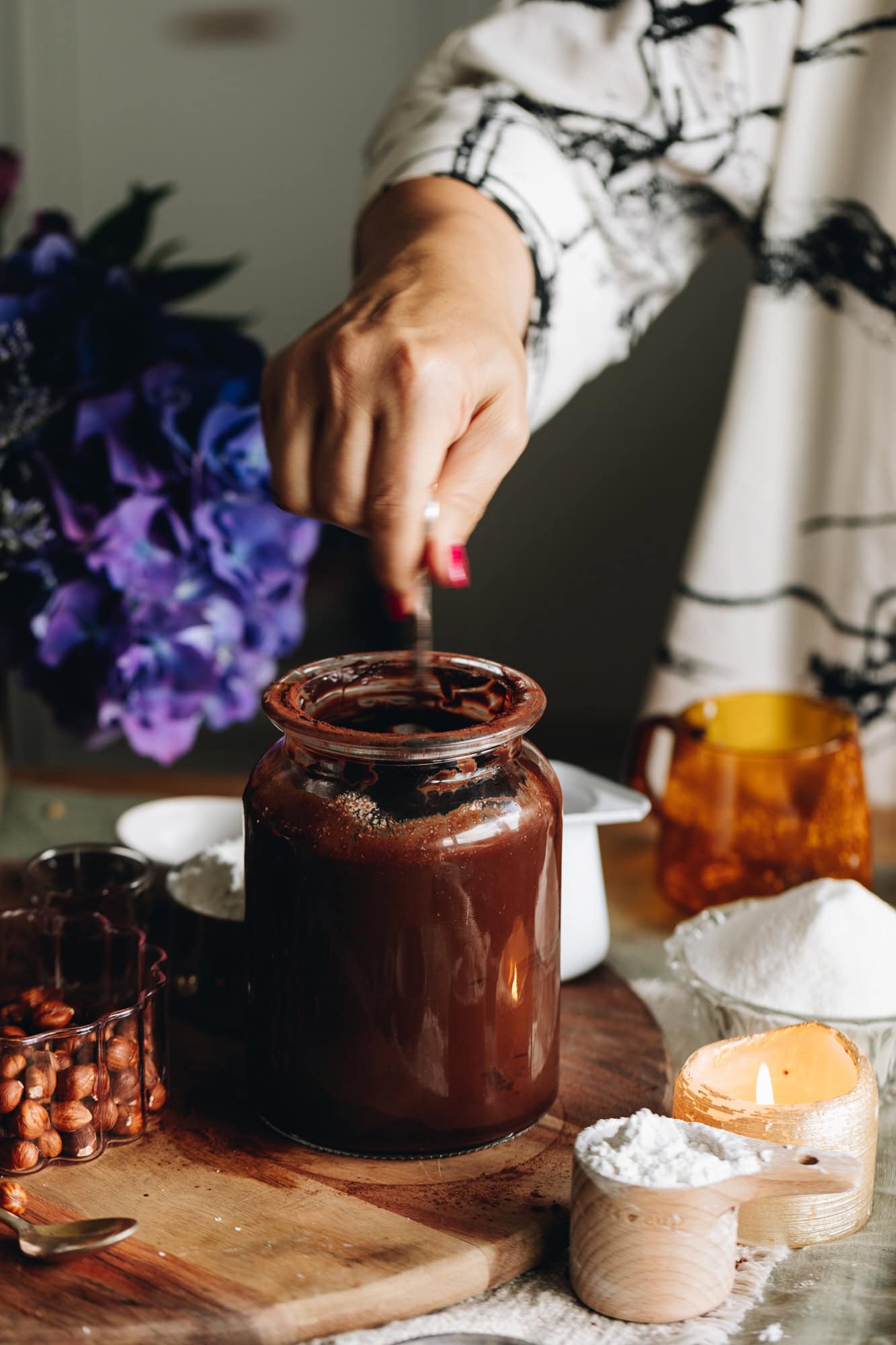 On a wooden board is a glass jar filled with a cocoa mixture. It is being stirred with a fork. Hazelnuts are also on the board and a candle burns next to it. Purple flowers are in the background.