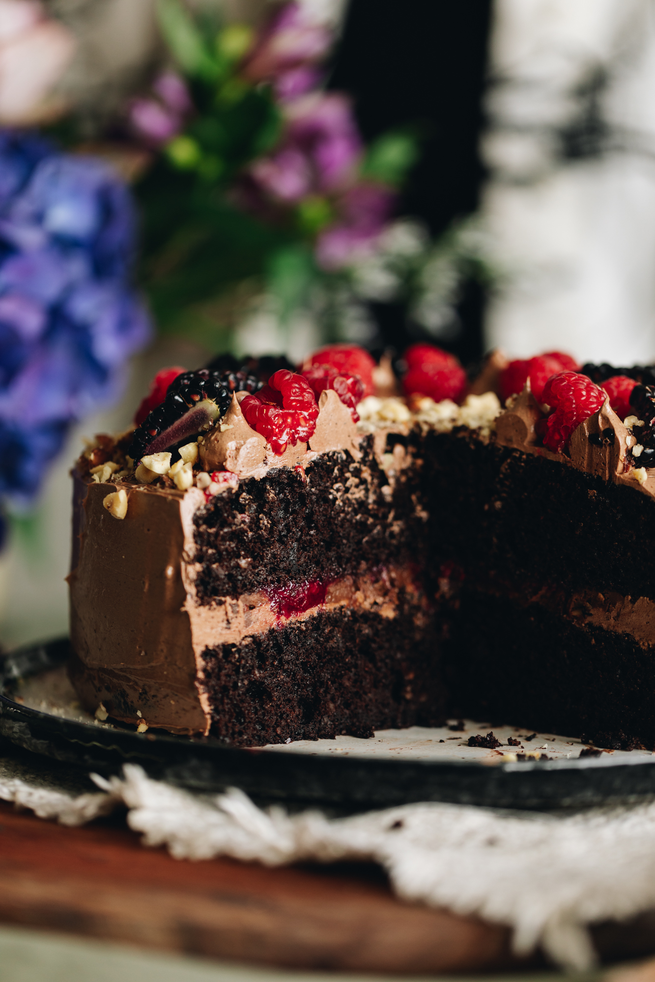 A black and white plate sits on a wooden board. On top is a gluten-free chocolate hazelnut cake which has piped buttercream and blackberries, hazelnut crumb and raspberries on top of it. It has been sliced open to reveal the insides of the cake showing the buttercream and boysenberry centre.