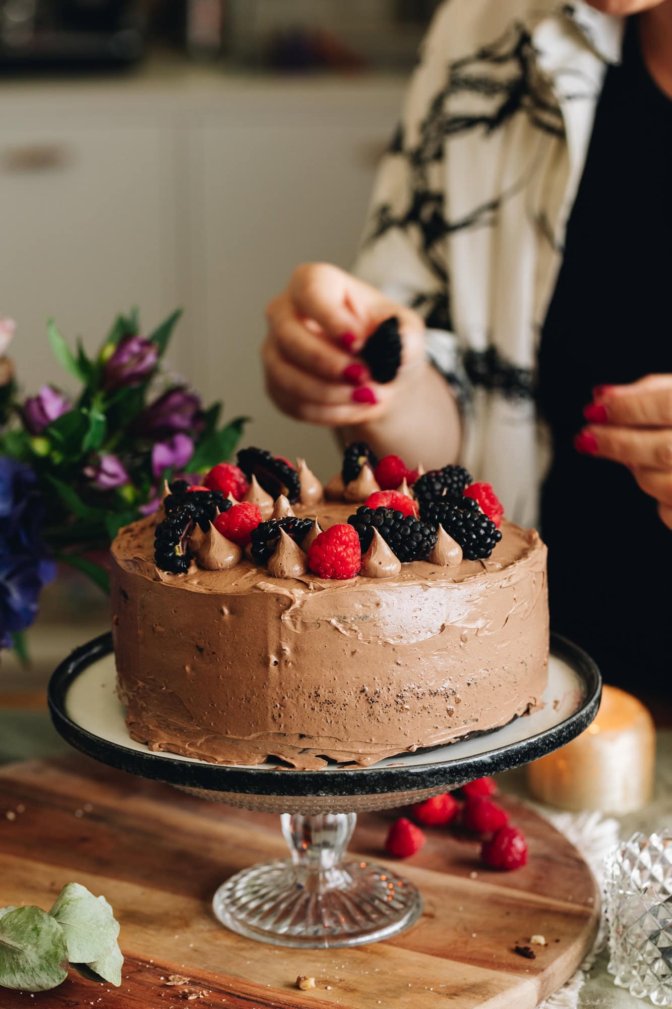 A glass cake stand has a black and white plate on it. On top is a gluten-free chocolate hazelnut cake which has piped buttercream and blackberries and raspberries on top of it.