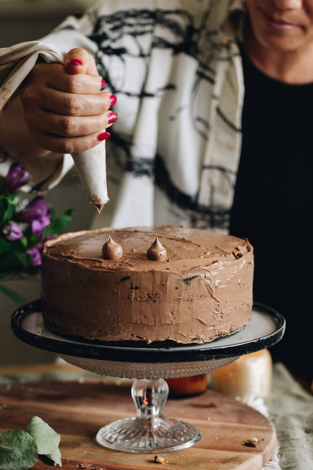 A glass cake stand has a black and white plate on it. On top is a gluten-free chocolate hazelnut cake which Naomi Toilalo is piping small blobs of buttercream on top.