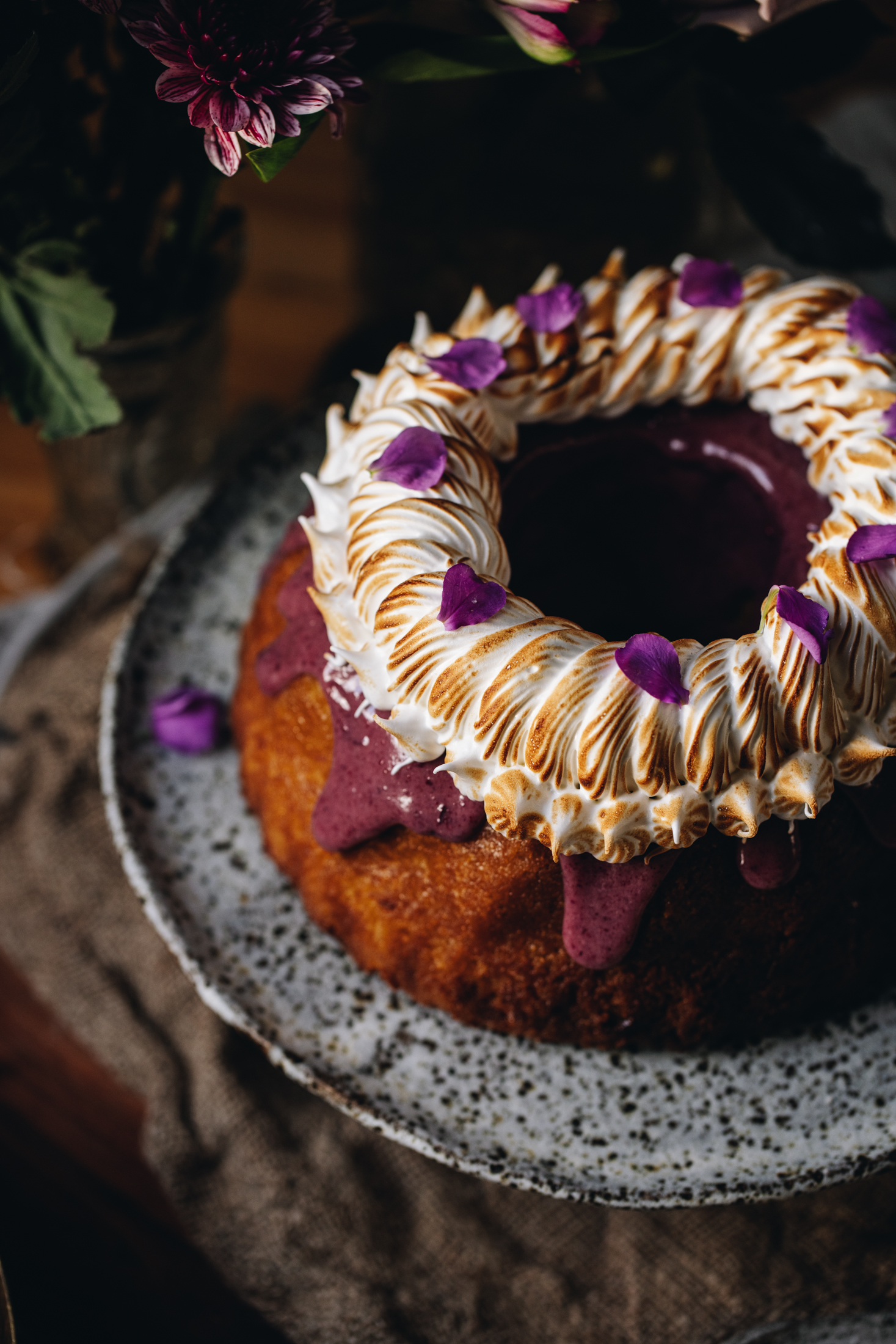 A top shot shows, a ceramic cake stand sitting on natural fabric with a lemon syrup bundt cake on top of it. It is decorated with a boysenberry ganache dripping down the sides and is topped with piped meringue that has been torched. There are flowers behind it.