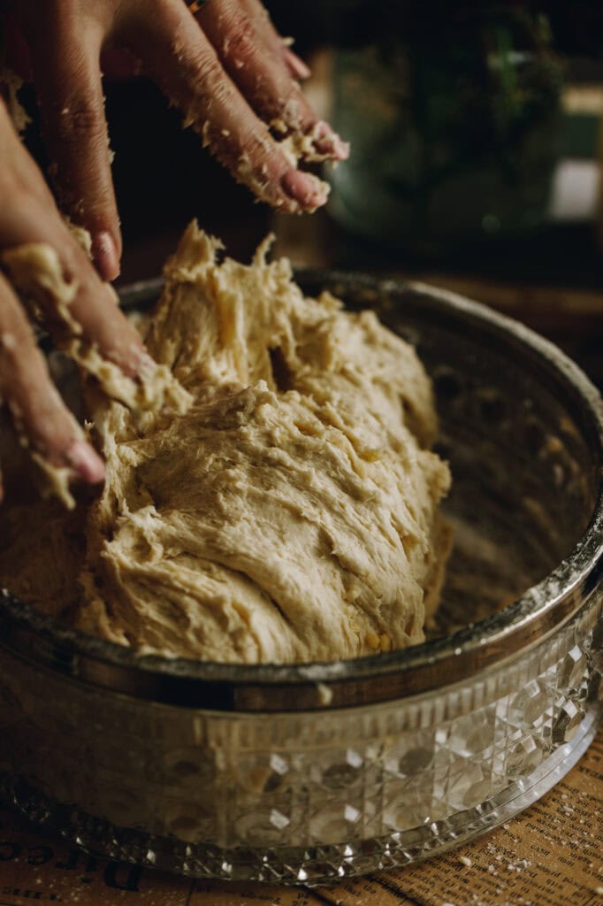 The dough is sticky in a glass bowl with a silver trim. Hands are working the dough with sticky dough on the fingers.
