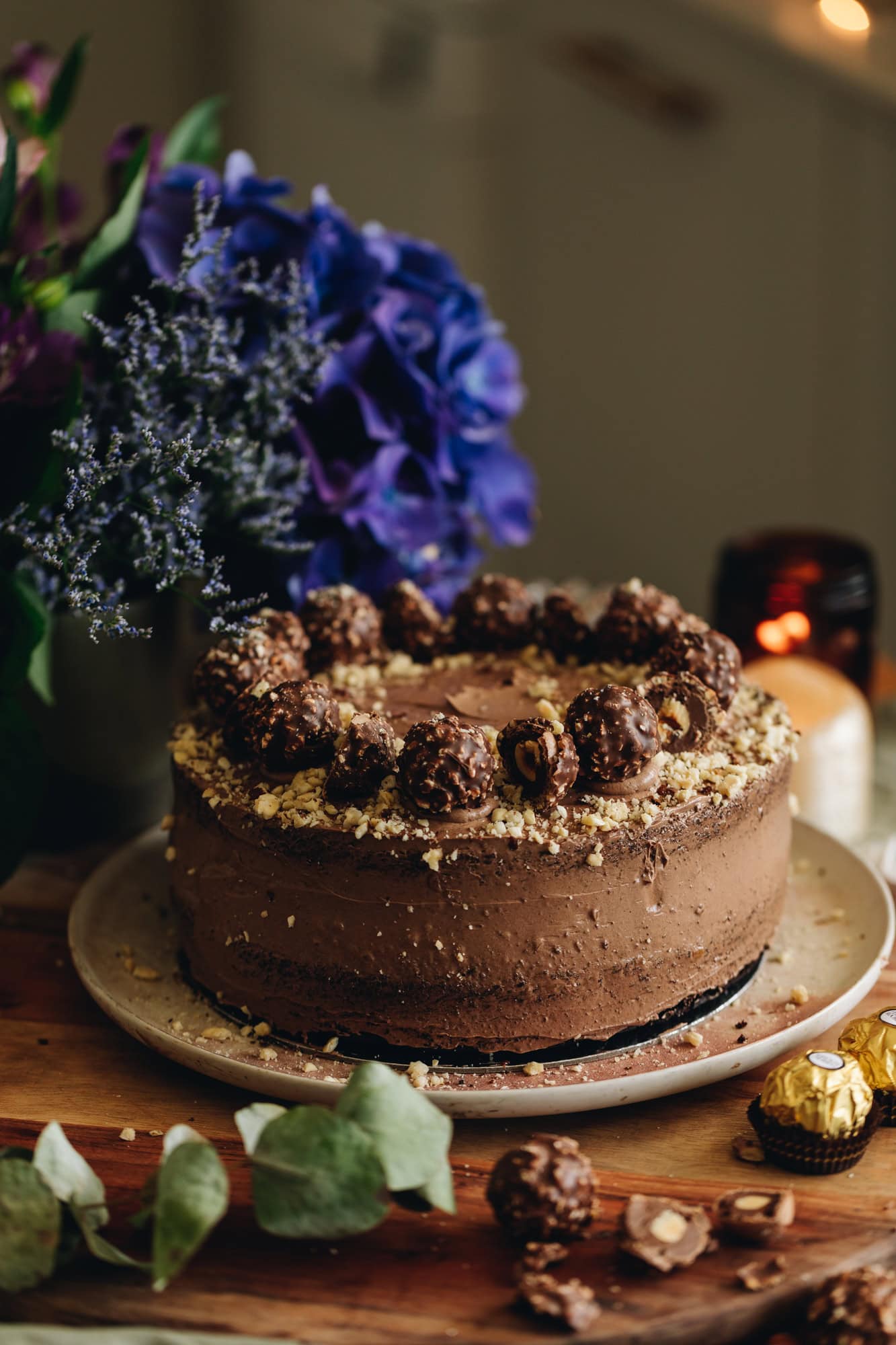 A whole chocolate hazelnut cake is sitting on a pink plate on a wooden table. It is covered in chocolate frosting with chopped hazelnuts and forerro rocher chocolates on top. It has purple flowers in the background. 