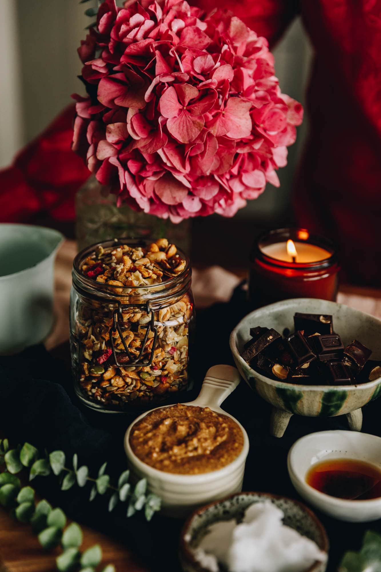 Ingredients are in different vintage bowls and are sitting on a wooden board on top of wooden table. There is muesli, peanut butter, vanilla and chocolate in view. There is a large pink hydrangea in the background and an amber candle burning next to it. 