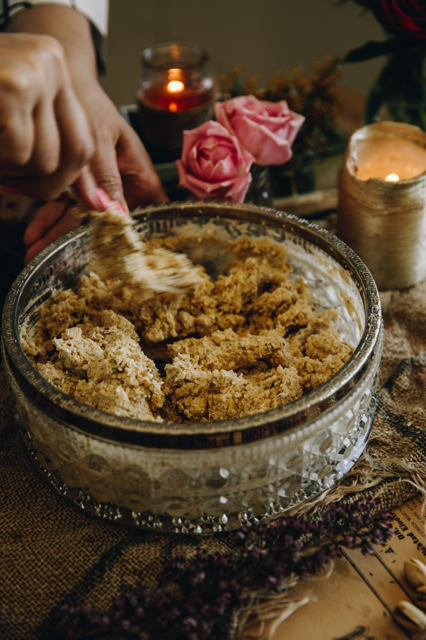 A fork is mixing the pistachio and chocolate cookie dough in a vintage bowl. It is on a wooden board with flowers and candles in the background.