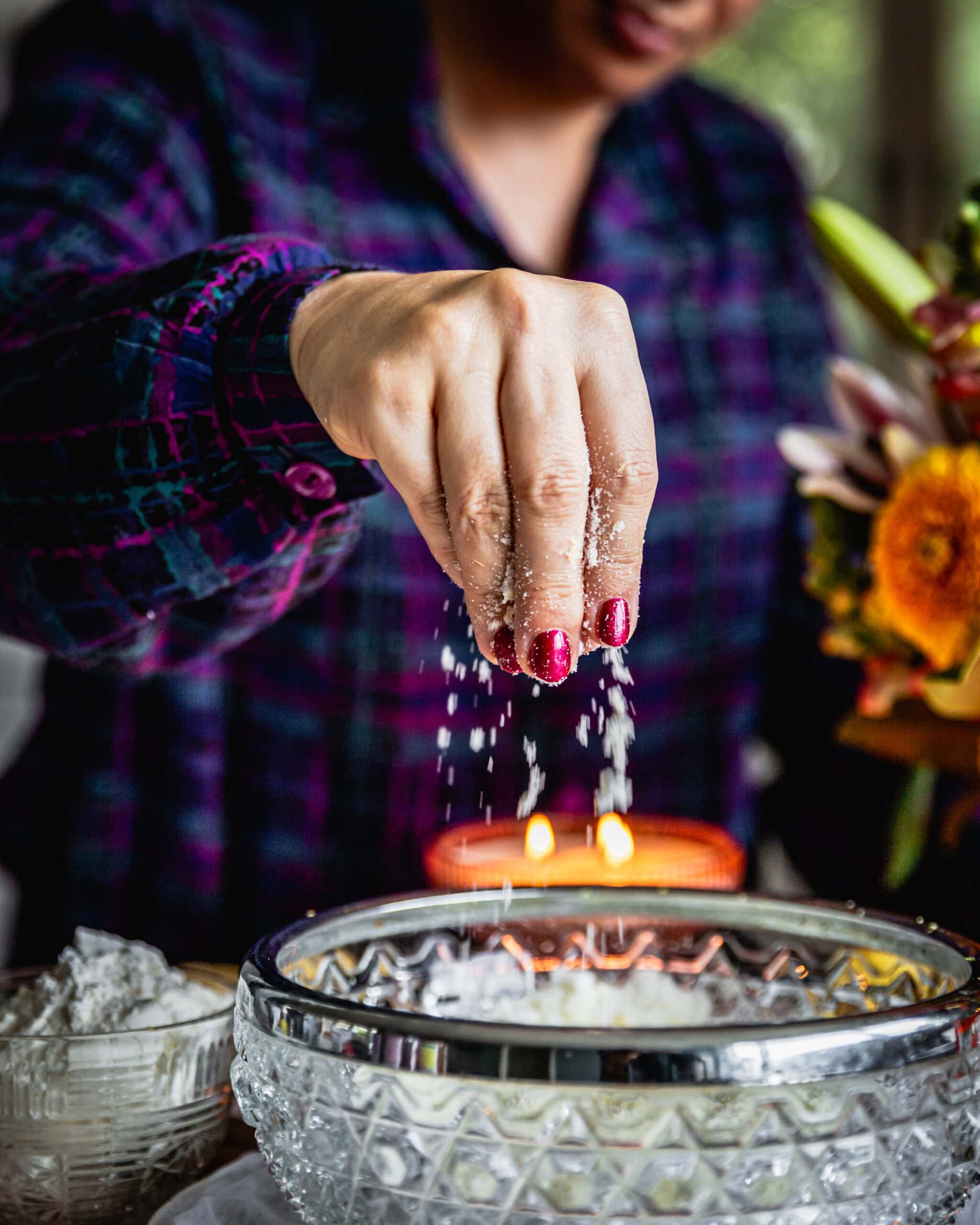 Naomi Toilalo is rubbing together sugar and lemon zest as she lifts it out of a glass vintage bowl. The action shot show the sugar sprinkling in to the bowl. 