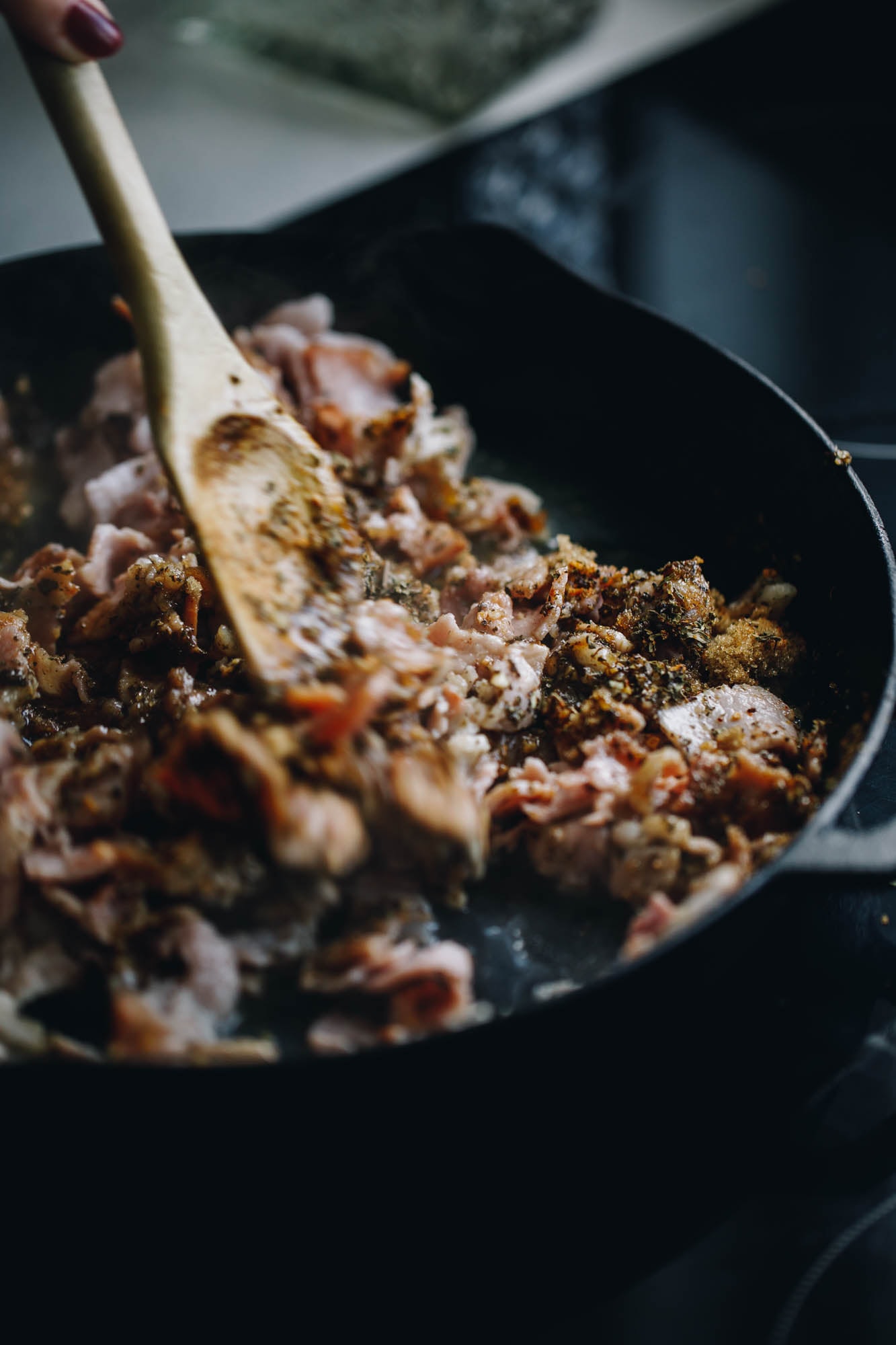 In a black cast iron pan is bacon with herbs frying. A wooden spoon is mixing it up.