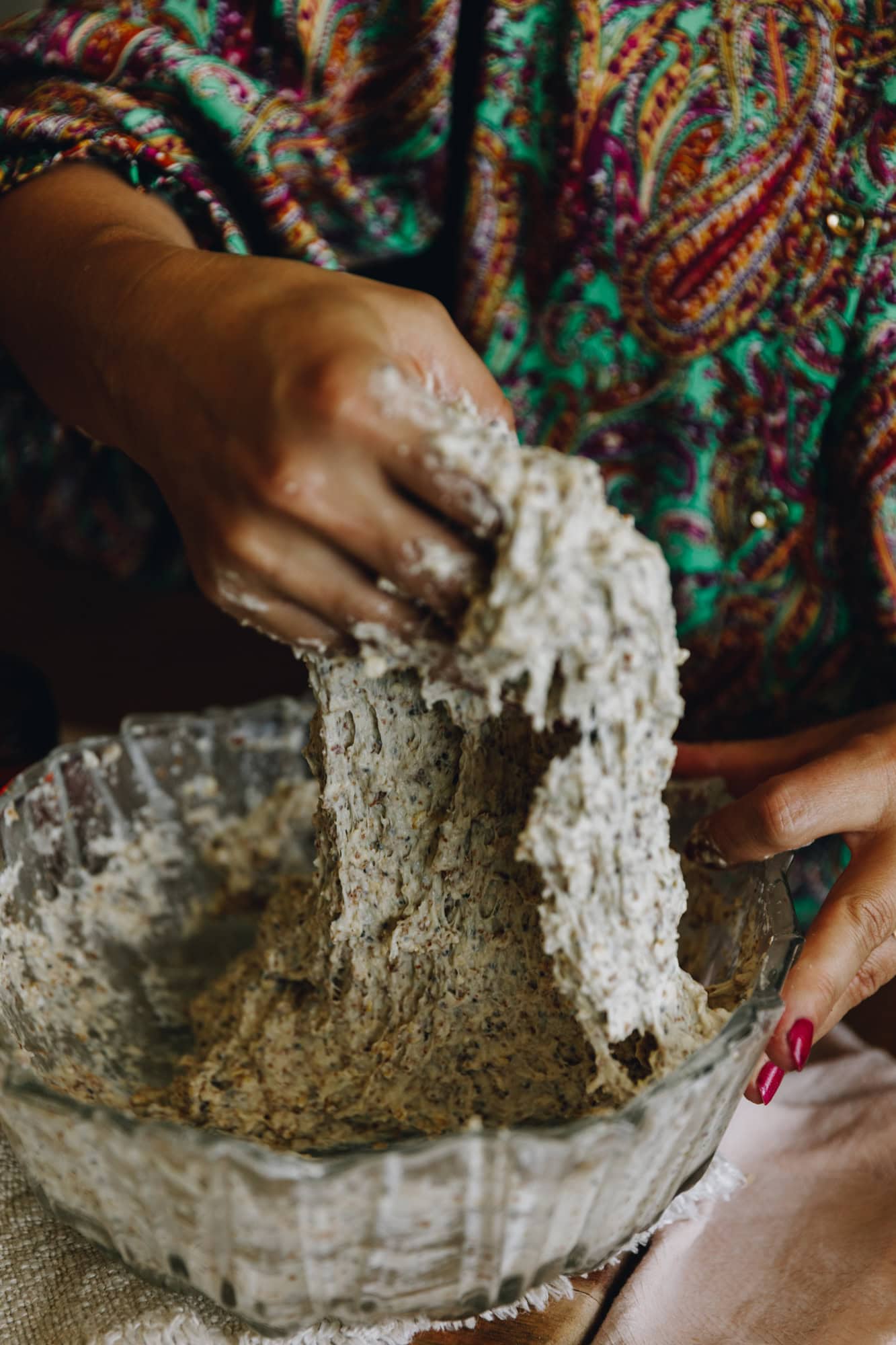 A hand with a colourful jumpsuit is gathering the dough of the no-seeded oat bread beginning to stretch and fold the dough. The dough is being lifted high in the air. 