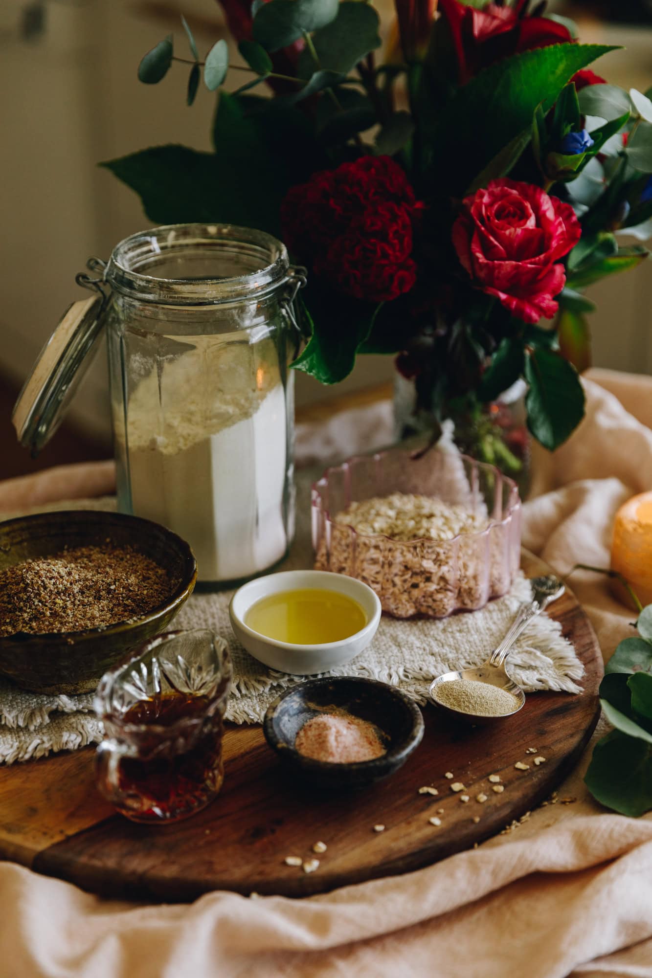 The ingredients for the no-knead-seeded loaf 
are in different vintage bowls and are sitting on a wooden board. A amber candle and pink flowers are behind the ingredients.