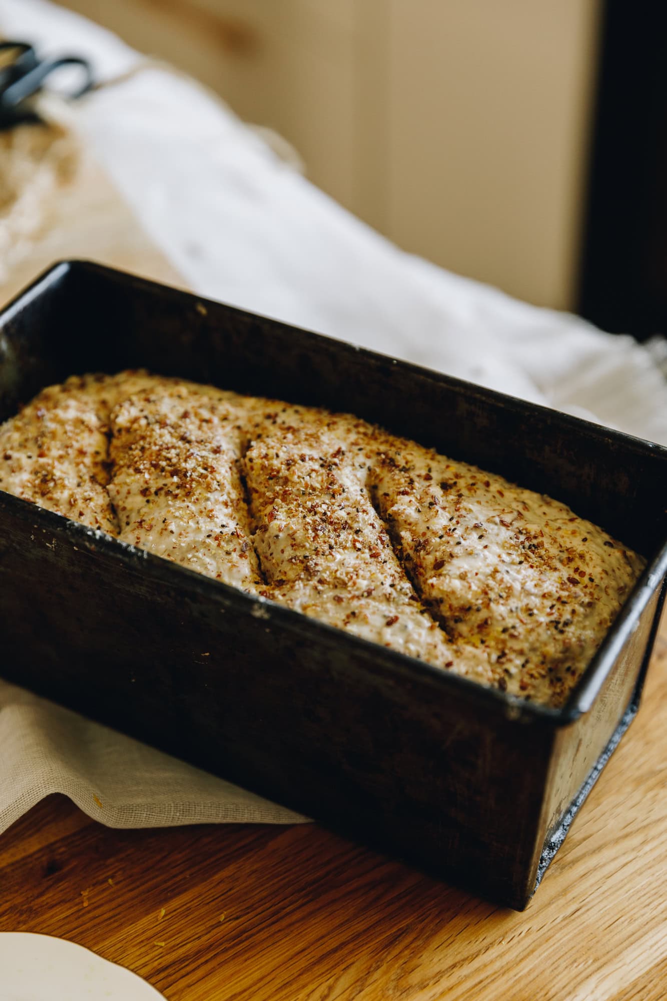 A no-knead seeded loaf dough is in a loaf tin unbaked on a wooden table. There are four slits on top of the bread and it has sprinkles of seeds on top.
