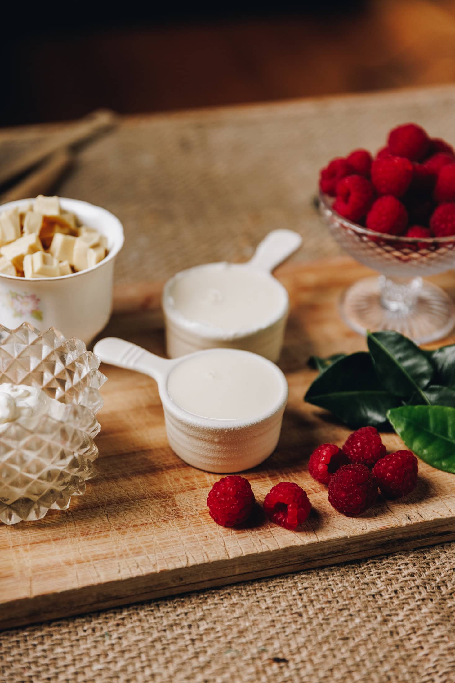 On a wooden board are fresh raspberries, lime leaf and cream and white chocolate in vintage glasses and cups.