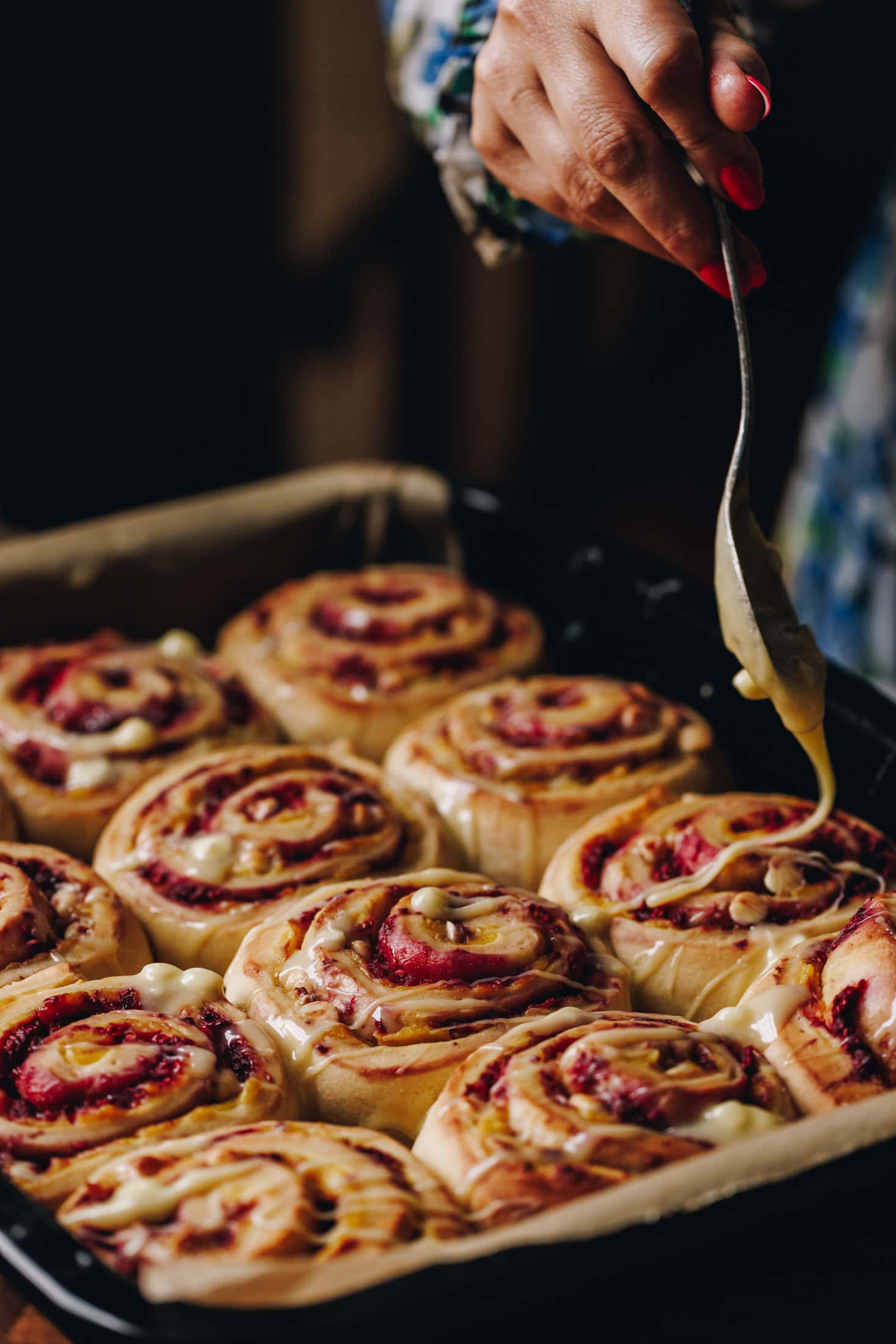 A black tray lined with baking paper is filled with baked custard and raspberry scrolls are having melted white chocolate drizzled on top of them with a fork by Naomi.