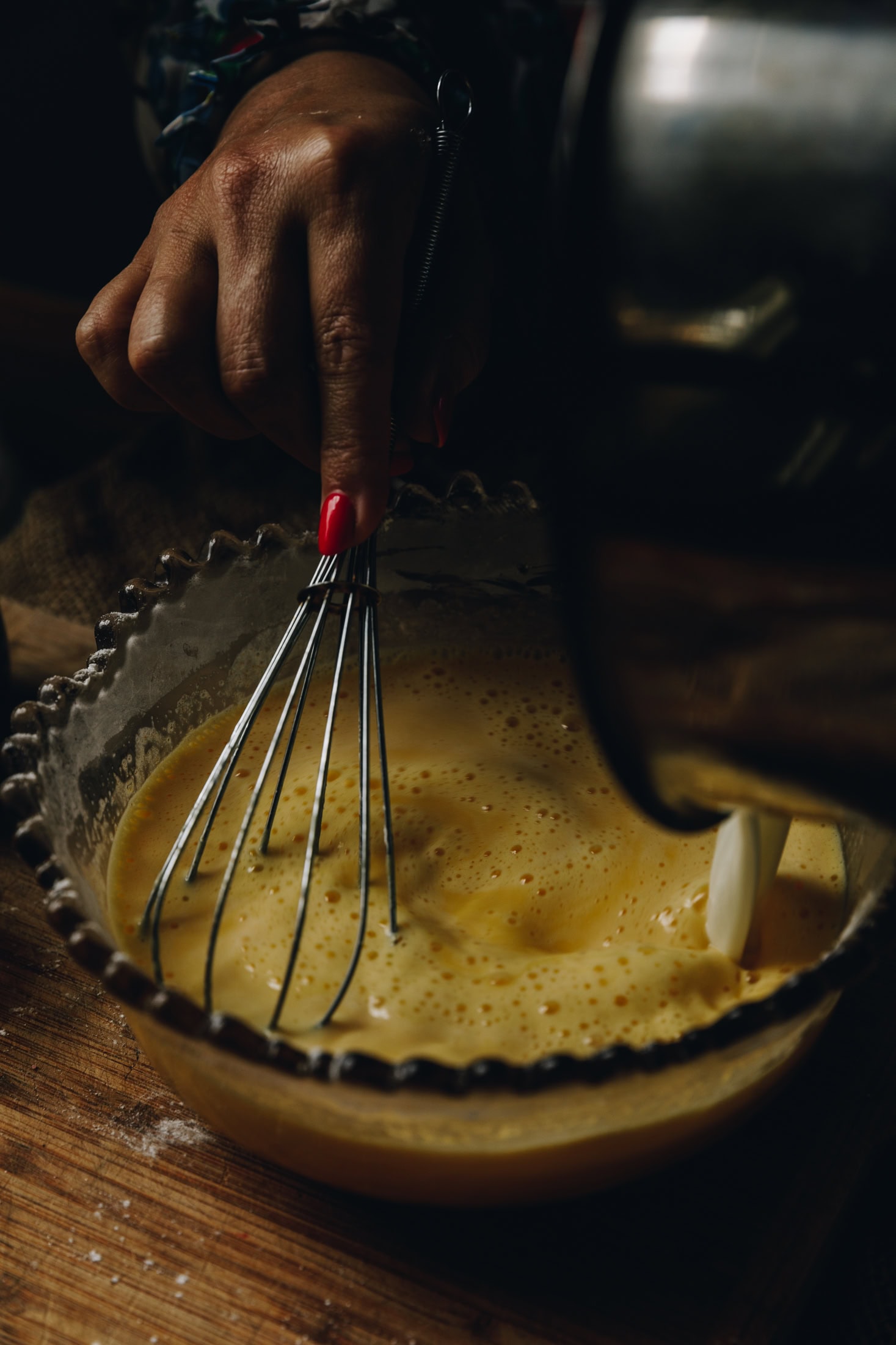 On a wooden table is a vintage bowl. Inside are eggs being whipped with a whisk as milk is being poured from a pot.