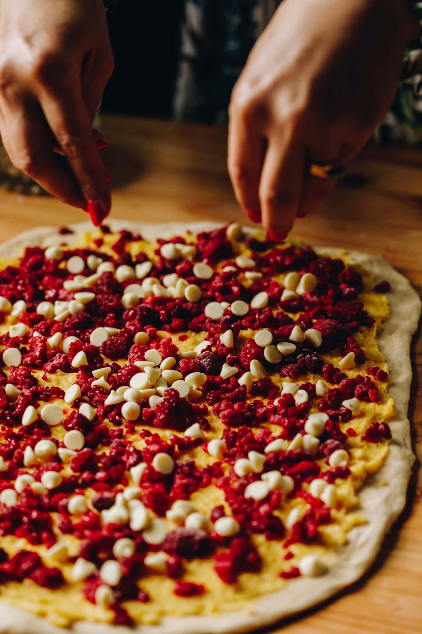 On a wooden table is the rolled out custard and raspberry scroll dough with the custard,raspberries and white chocolate drops all over it.