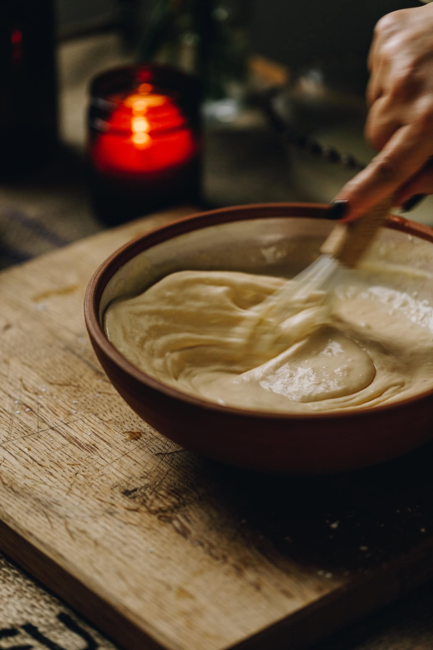 A ceramic bowl is on a wooden board, in the bowl is a lemon custard that is being stirred. An amber candle burns in the background.