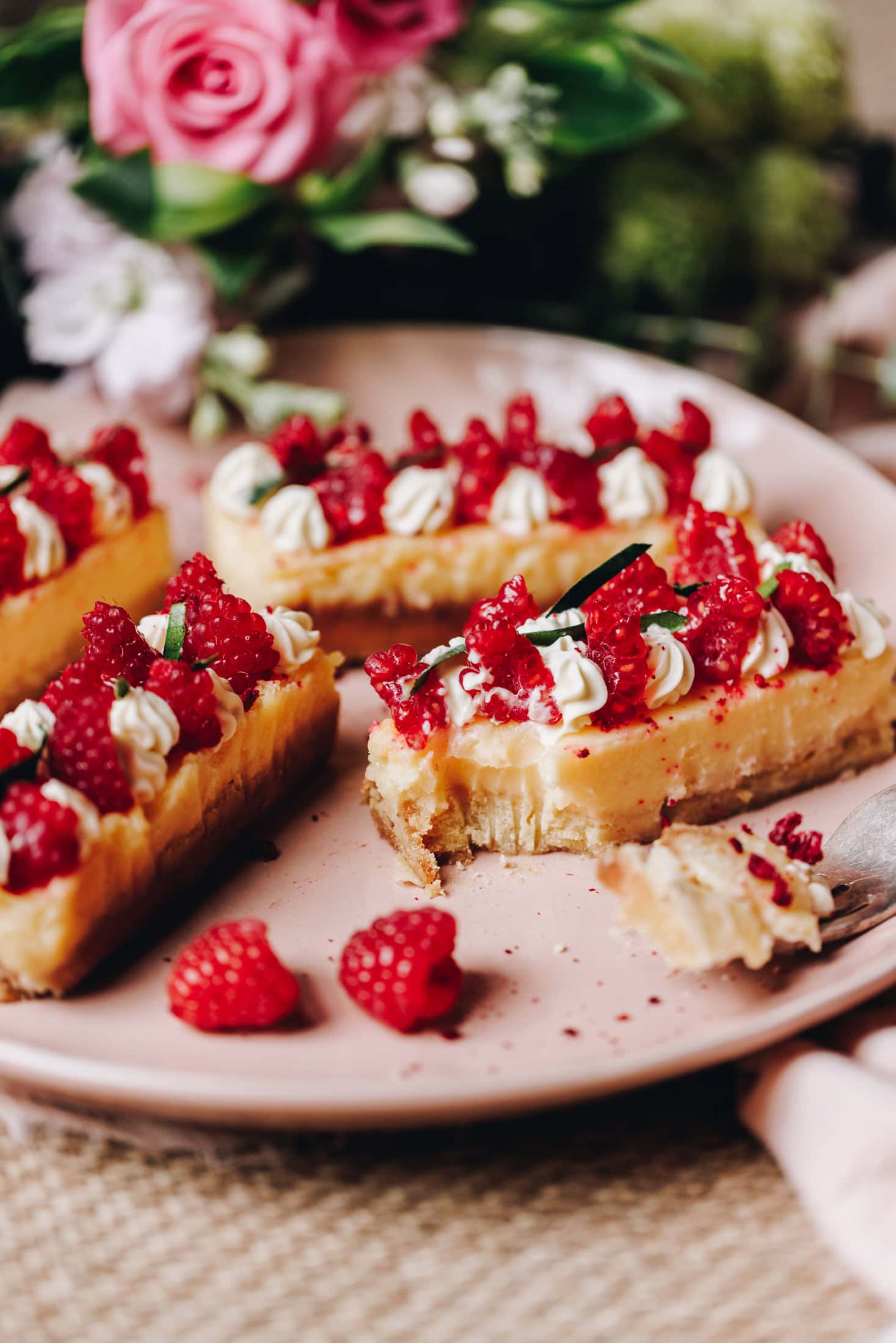 On a pink plate is lemon bars that are decorated with piped whipped cream, slices of fresh raspberries and slivers of lime leaves. Once of the slices have had a piece taken out of the side of it. Pink roses are in the background.