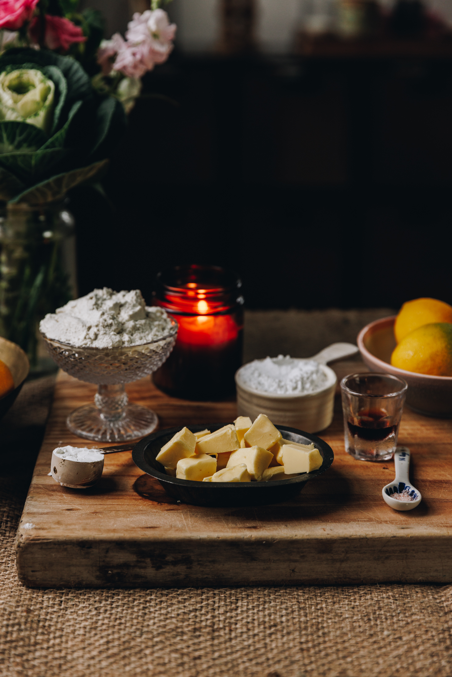 Ingredients of butter, icing sugar, vanilla and lemons are in different vintage bowls and are sitting on a wooden board on top of wooden table. There an amber candle burning and a bunch of flowers in the background.