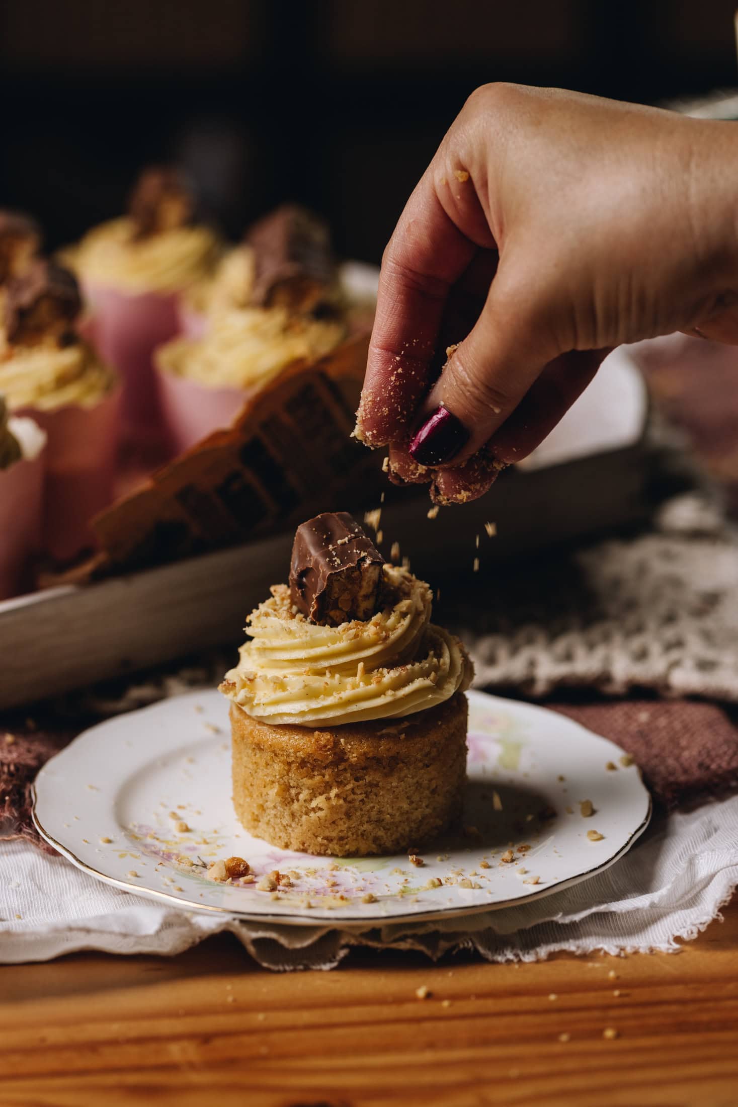 A hand is sprinkling over chopped nuts on to brown butter vanilla cupcakes with piped buttercream and a snickers slice on top.