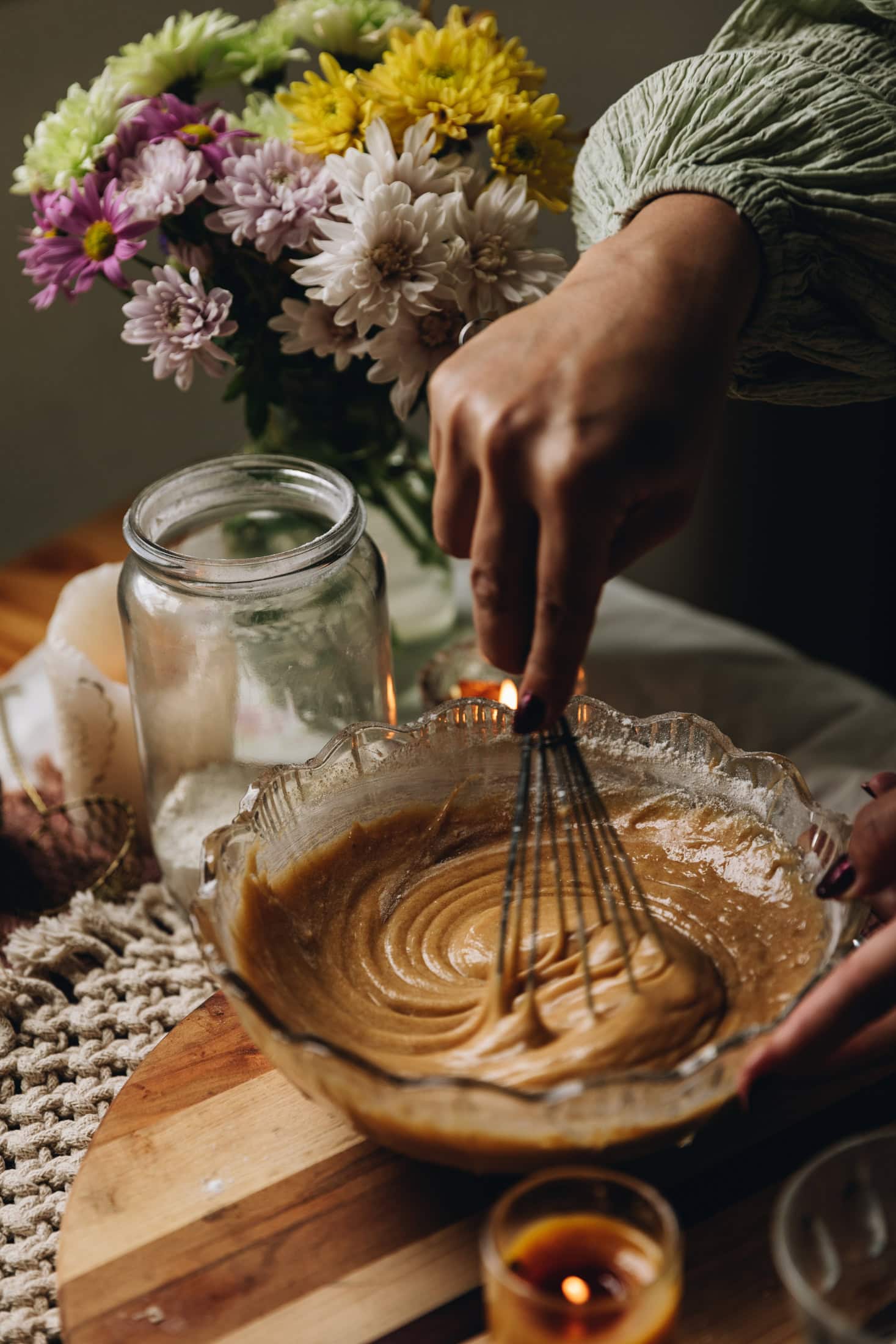 A glass bowl is sitting on top of a wooden board. A hand is stirring the unbaked vanilla cupcake mixture with a whisk.