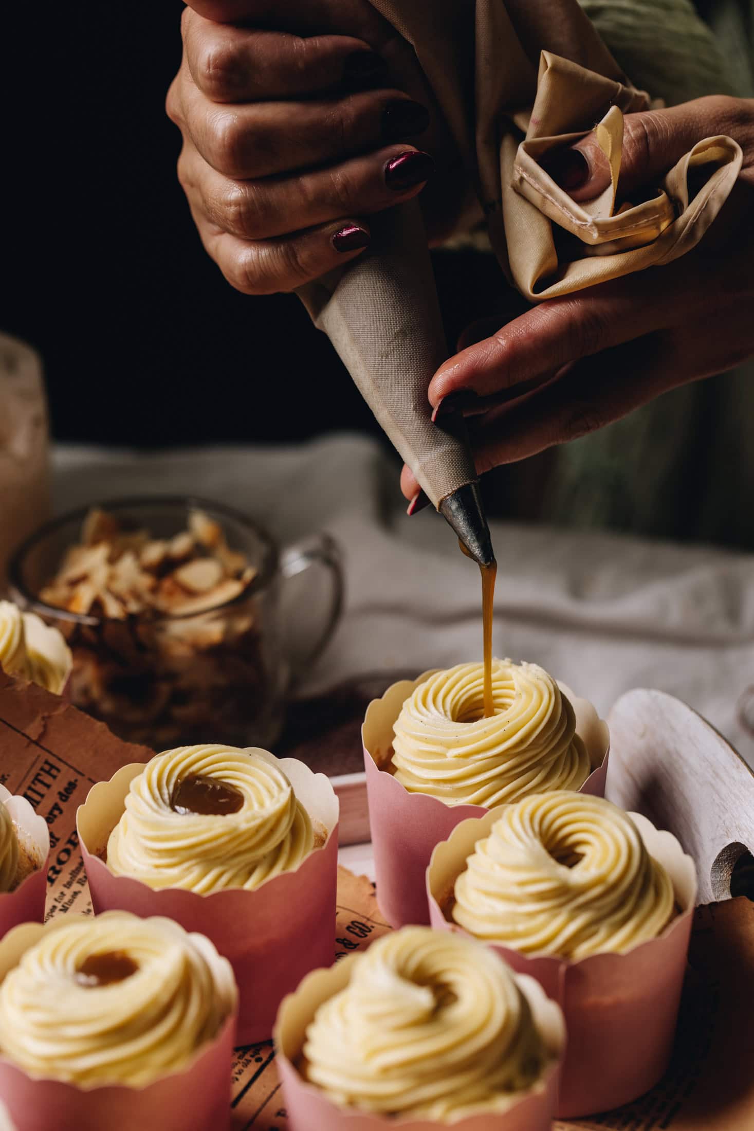A piping tip drizzling caramel in to freshly baked vanilla cupcake. Buttercream is seen on the top of other vanilla cupcakes in pink cardboard cases.