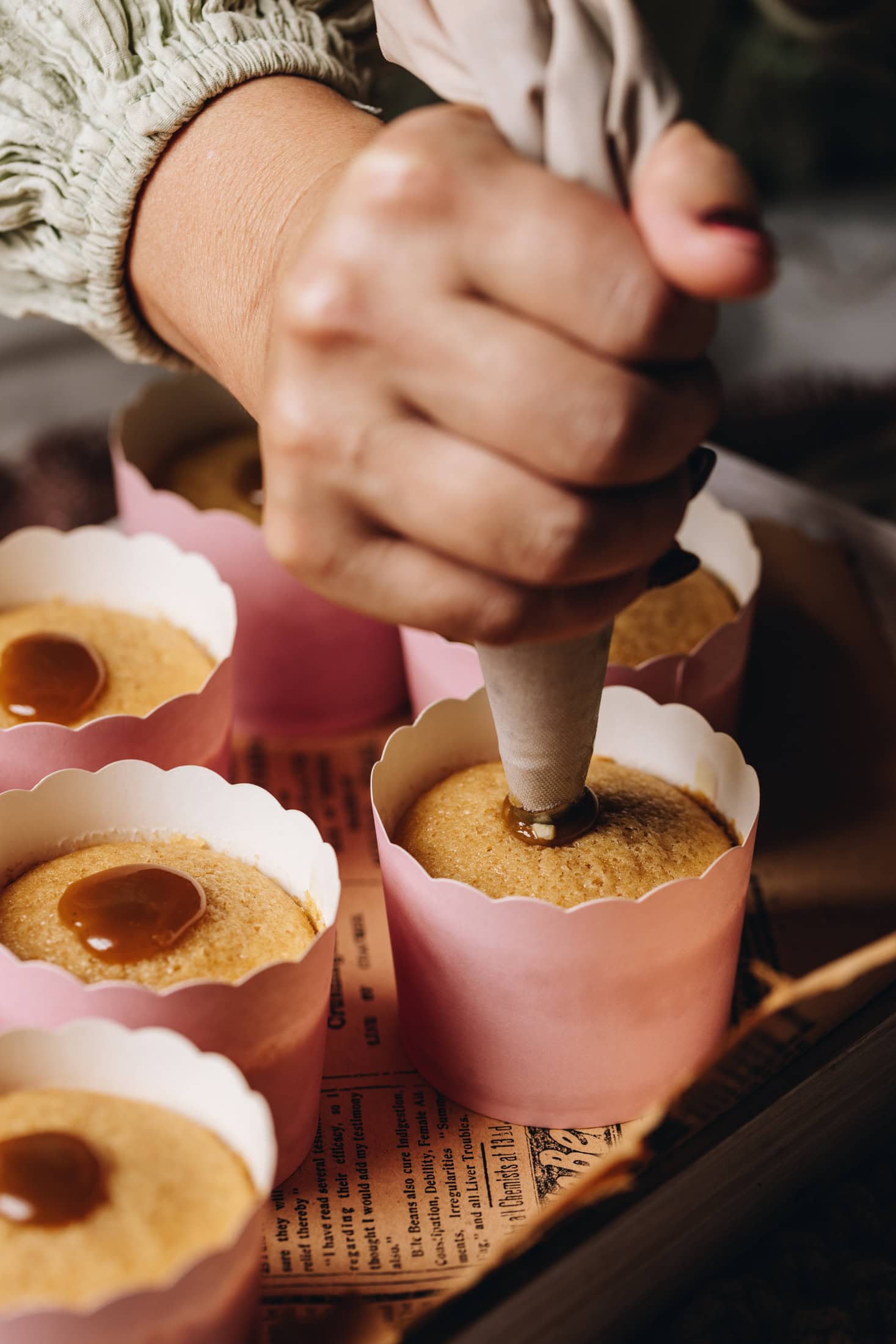 A piping tip is pierced into a freshly baked vanilla cupcake. Caramel is seen on the top of other vanilla cupcakes in pink cardboard cases.