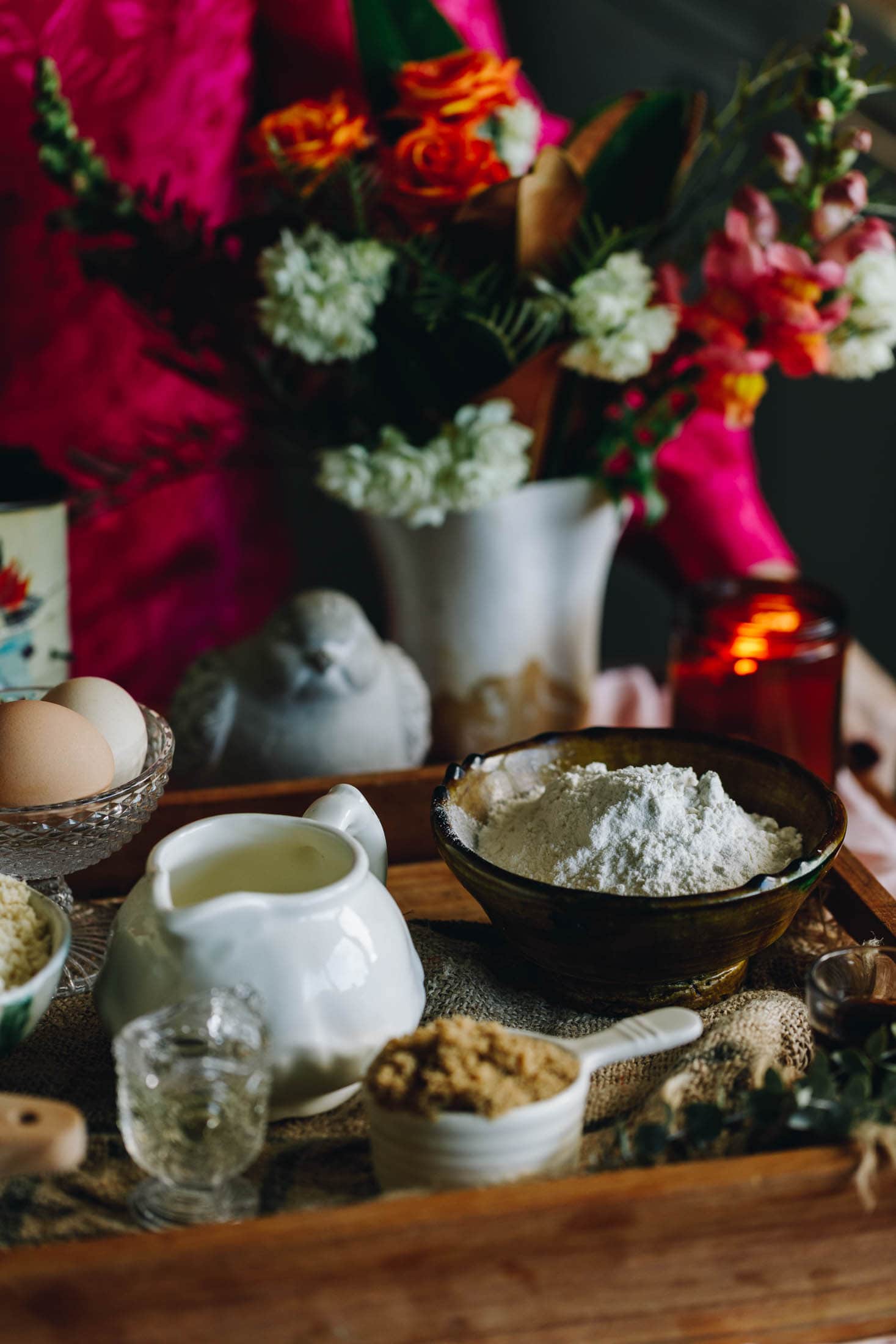 A wooden tray displays the ingredients for the Chocolate Cake with Biscoff Buttercream in beautiful jars, jugs and cups. There are eggs, flour and sugar in view. A bunch of flowers are in the background with a ceramic bird. 