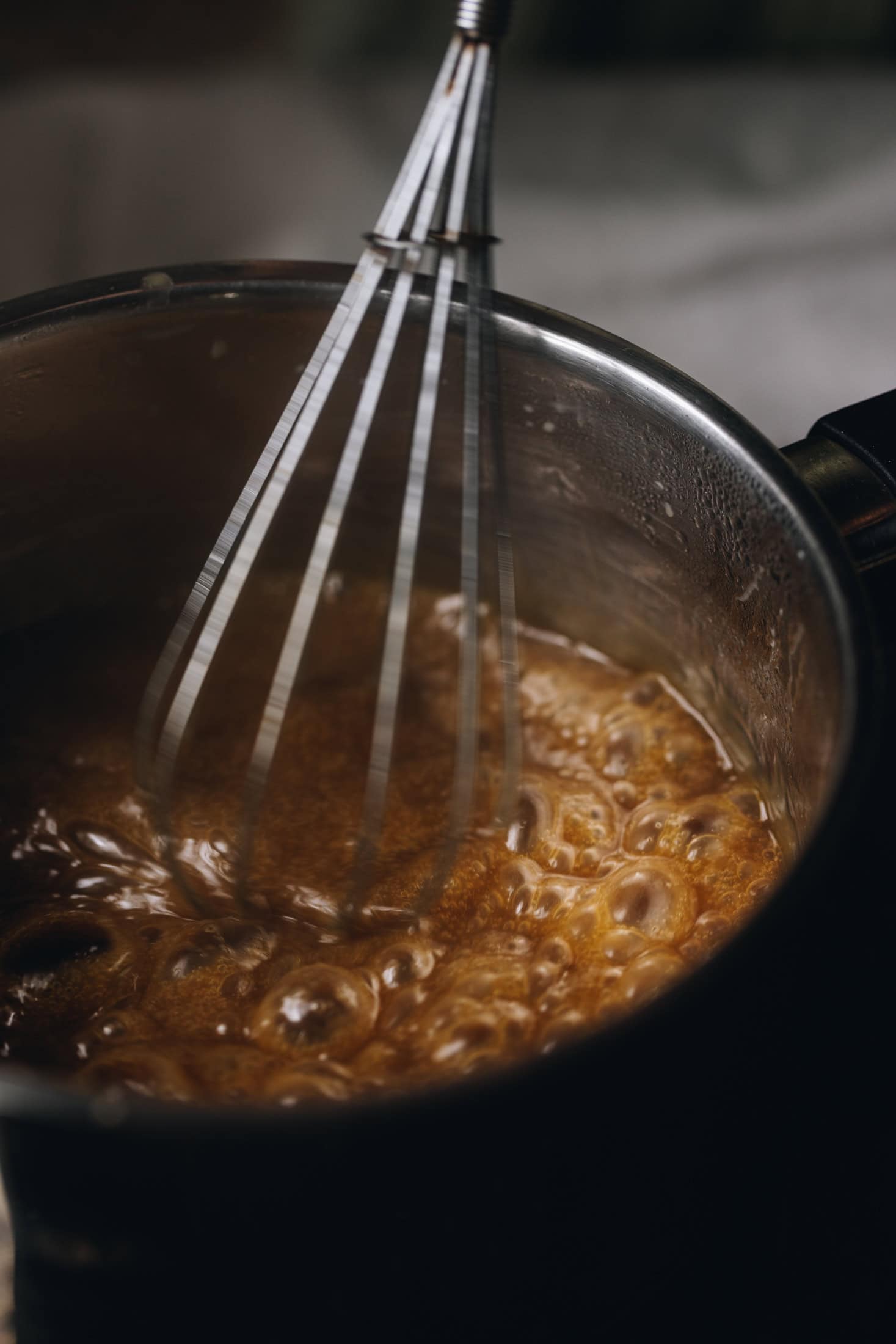 A silver pot has caramel boiling in it, it is deep in colour and is being stirred with a silver whisk.