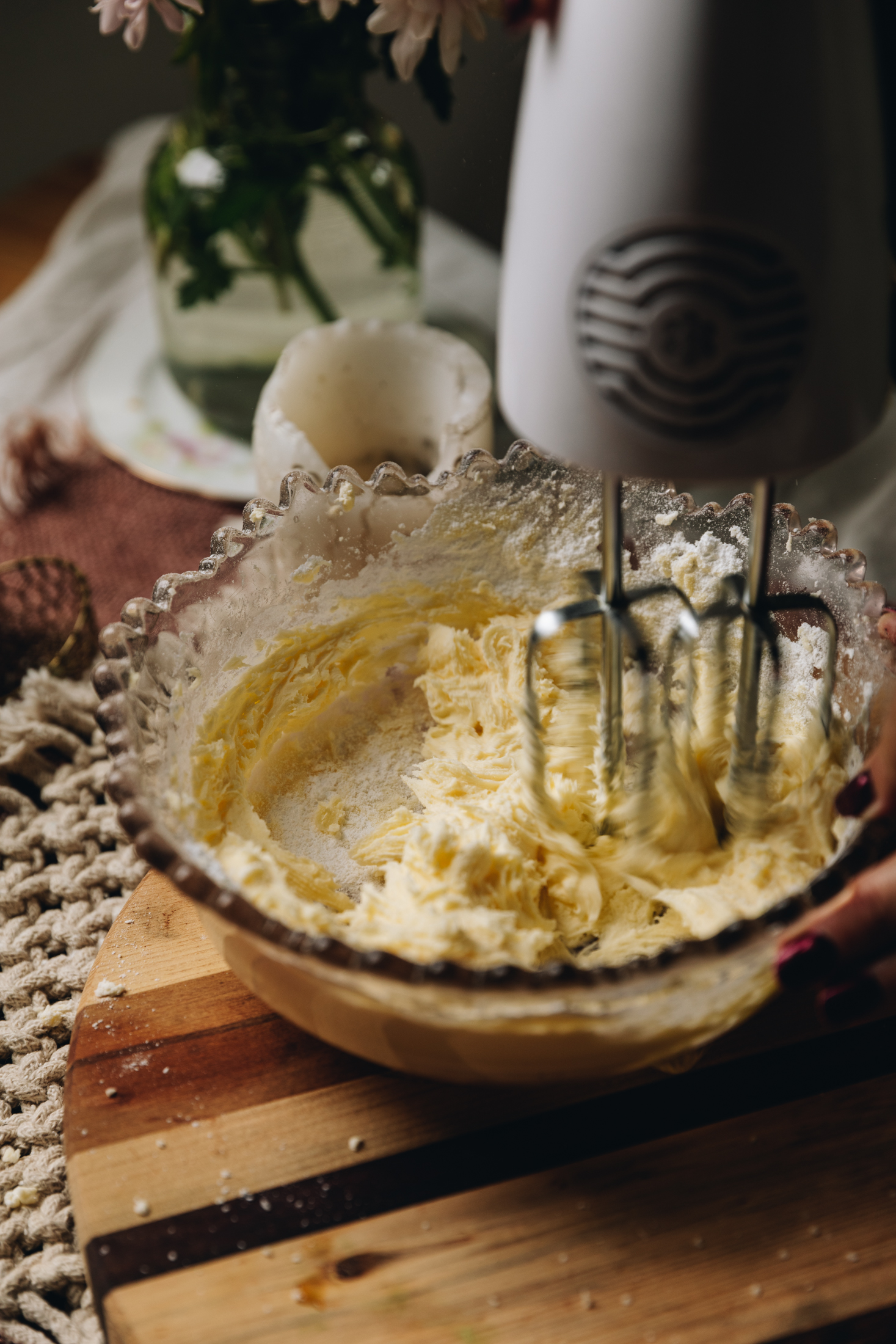 A vintage bowl with a crinkled edge is sitting on a wooden board. Inside is a cream cheese frosting that is being whipped with a hand mixer.