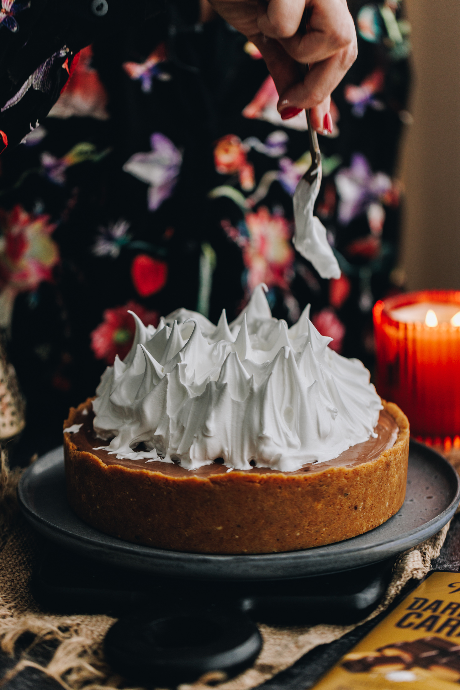 On a table sits a blue table with a s'mores cheesecake on it. It has a biscuit base and Naomi is decorating an Italian meringue on top. She is using a fork to make flame shapes with it. An orange candle burns in the background. 