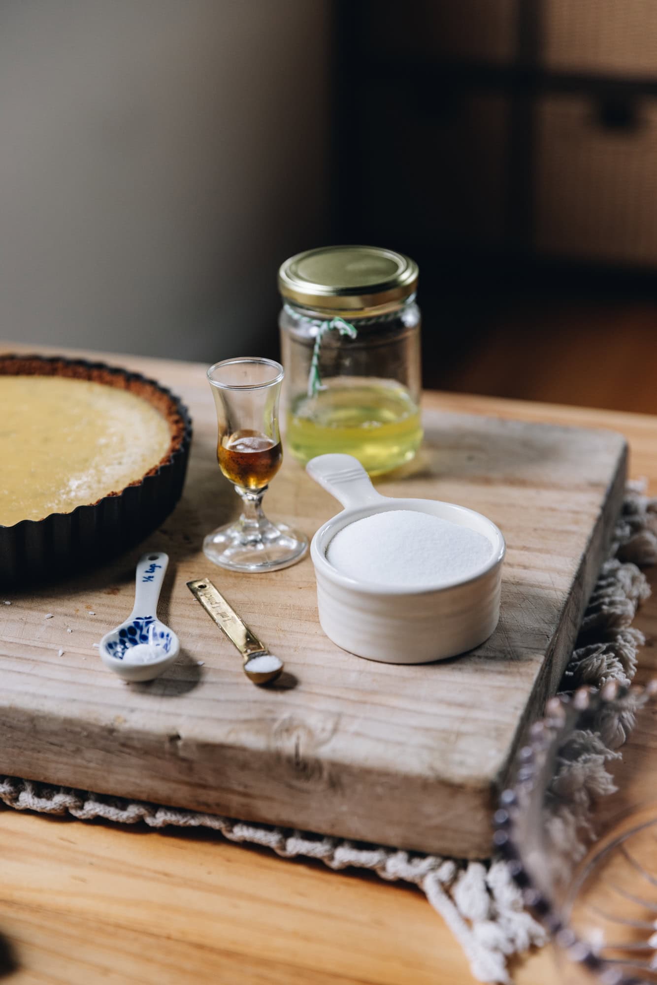 On a wooden board sits vintage cups and spoons that are holding the ingredients for the Italian meringue. A baked lime pie is on the side of the board. It sits on a cream weaved mat, on top of a wooden table. 