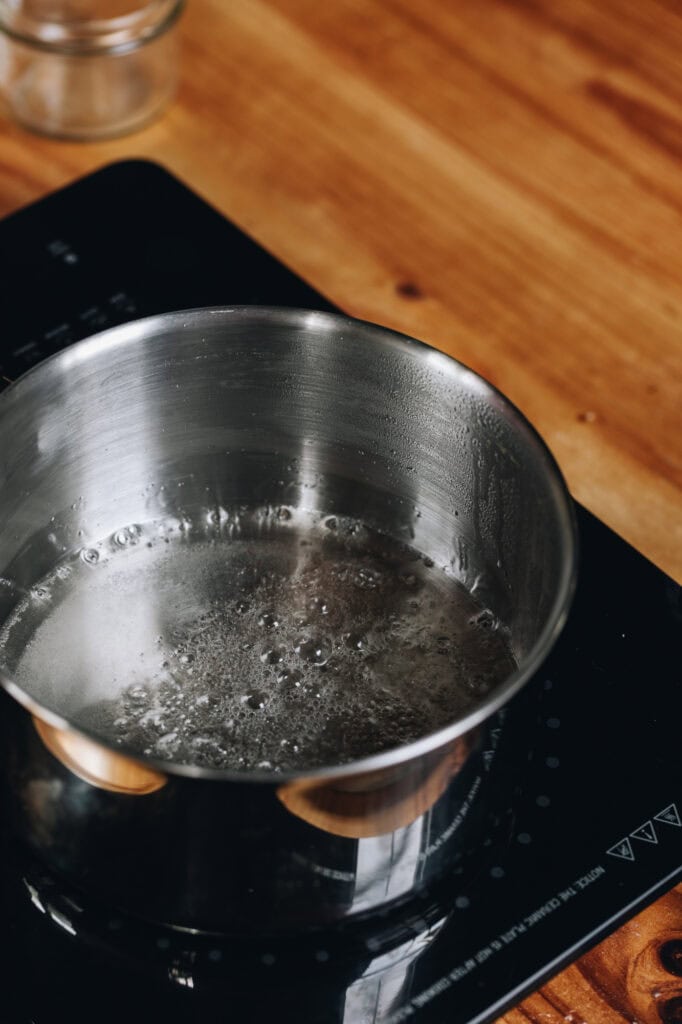 On a wooden table sits a black portable stove top. On it sits a silver pot with sugar syrup bubbling in it. 