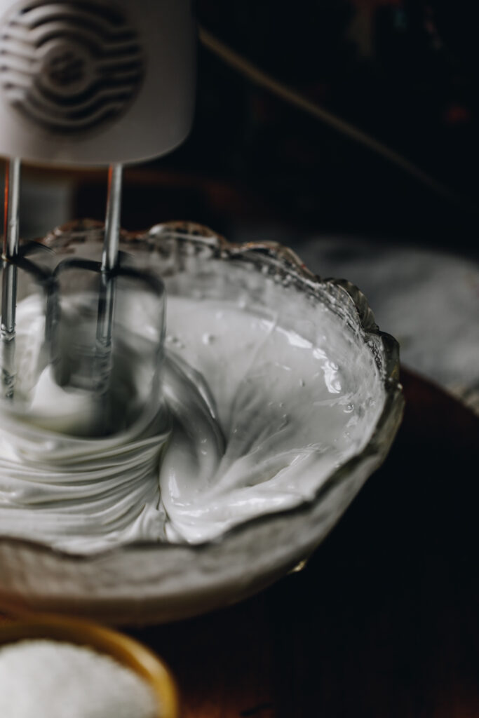 A glass bowl is filled with a meringue that is being whipped by a hand beater. It is glossy and the shot is moody. 