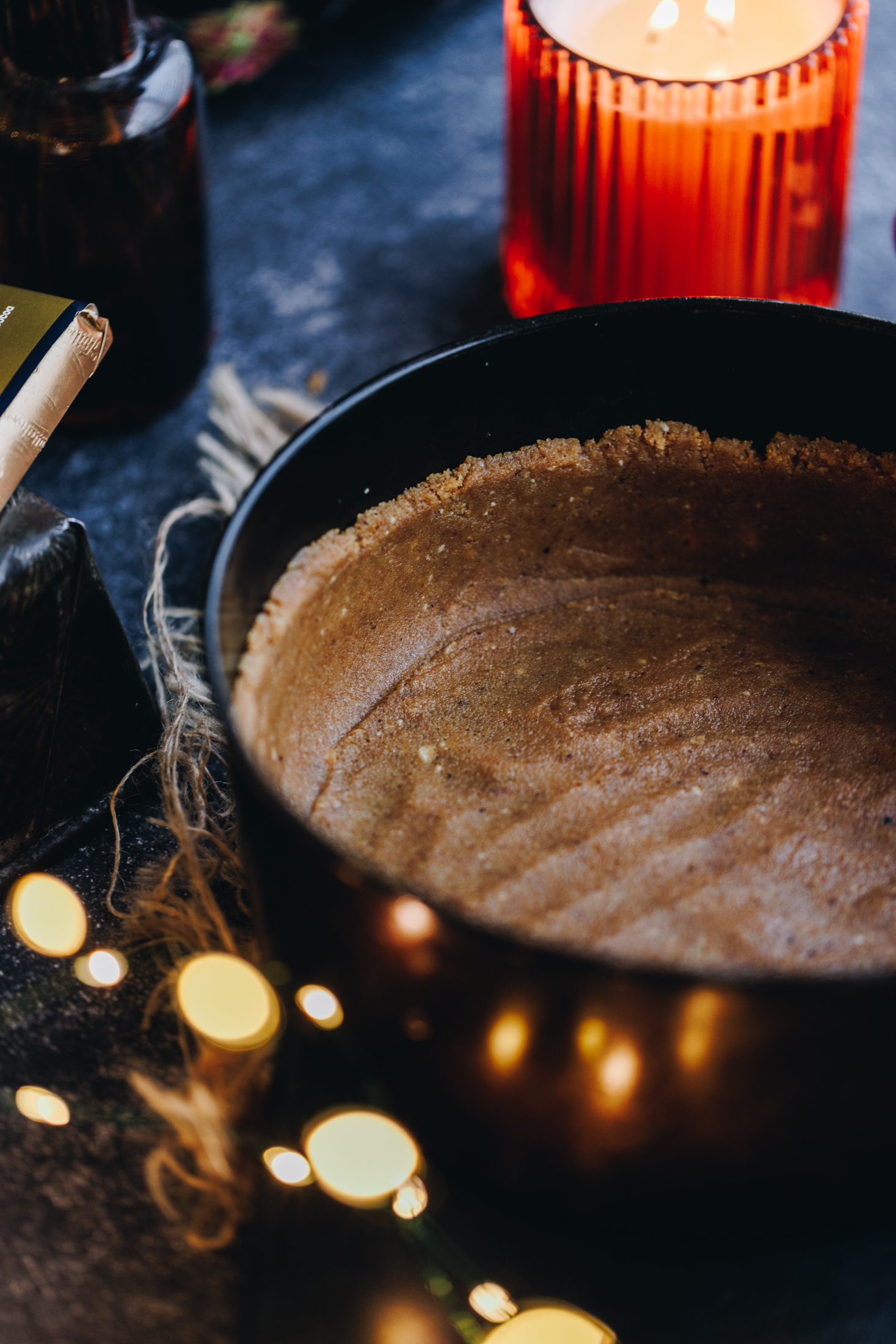 The biscuit base of the S'mores No-bake Chocolate Cheesecake has been pressed in to a black car tin. An orange candle burns in the background. 