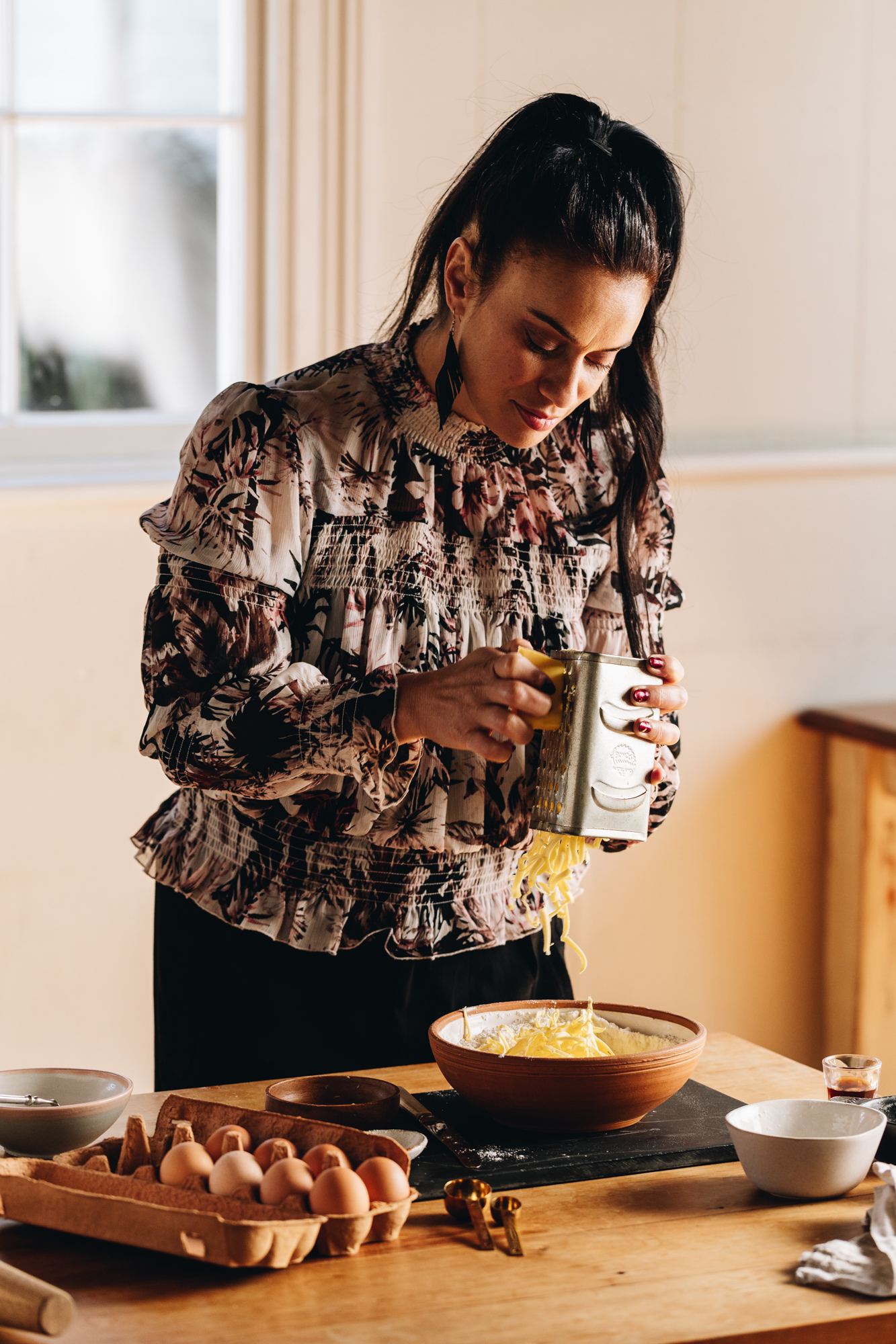 Naomi Toilalo is standing behind a wooden tabled. On the table is a black stone with a ceramic bowl on top. Naomi is grating butter in to the bowl. Next to her is a carton of eggs open, and a couple of white bowls and baking equipment.