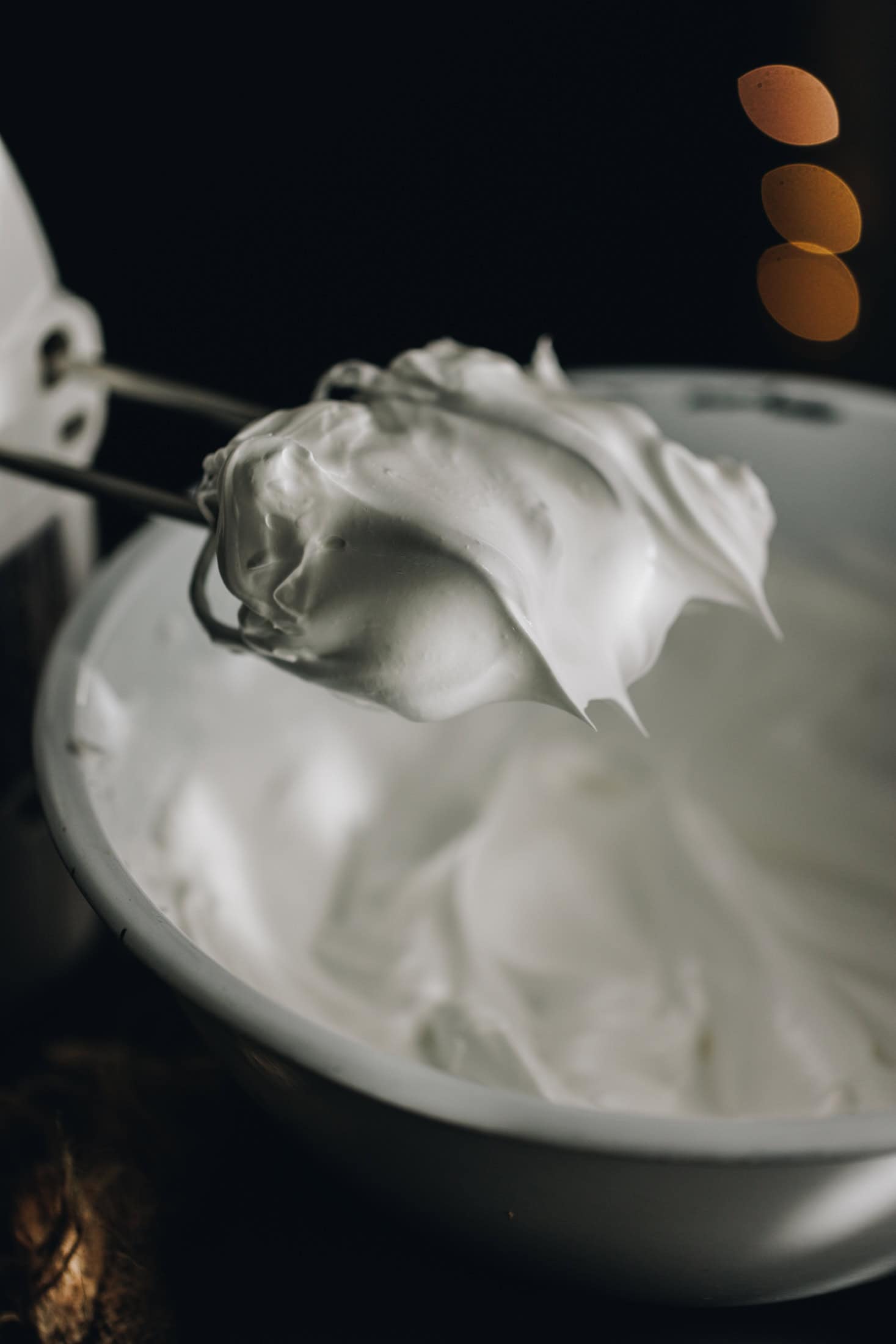 A meringue is in a white bowl and a hand mixer holds stiff peaks of Italian meringue at the tip of the beaters. 