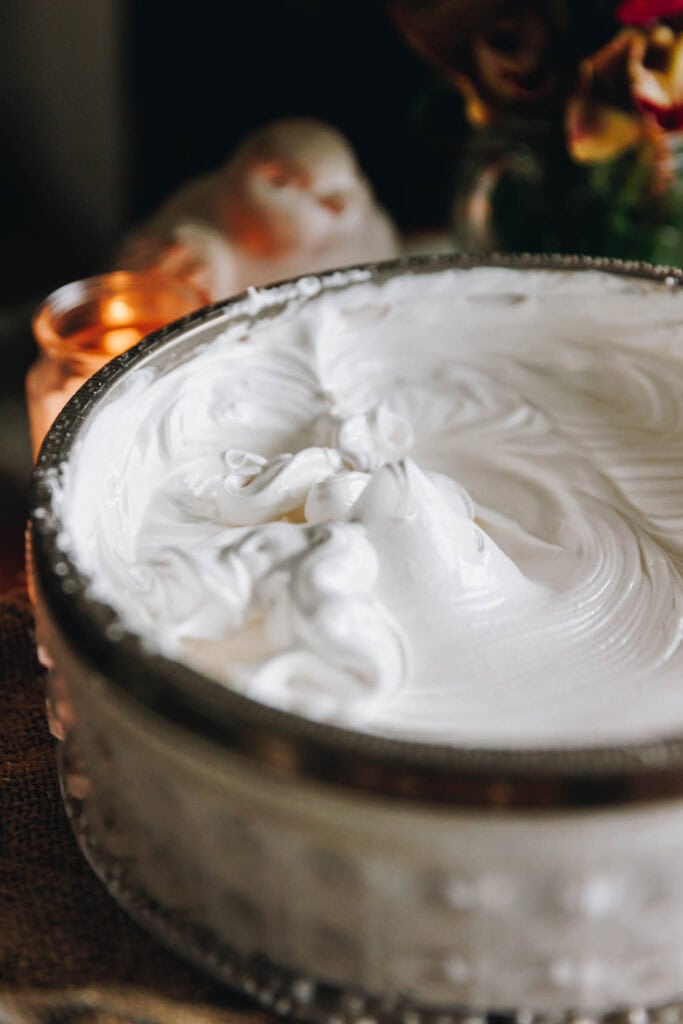 A glass bowl with a silver trim is filled to the brim with silky meringue that is white and fluffy and shiny. 