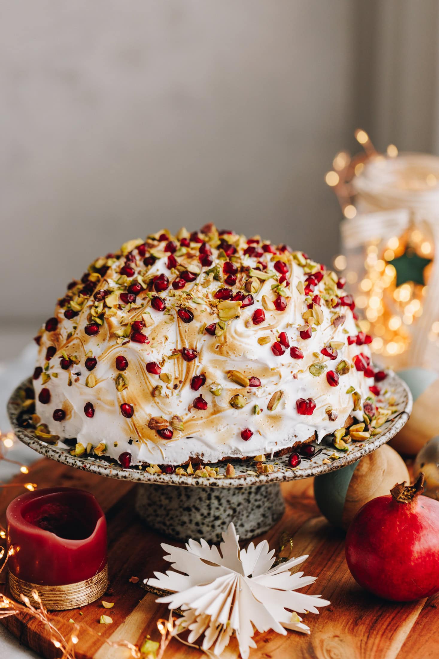 A cheat's ice cream Bombe Alaska is plated on a ceramic cake stand with meringue on top, chopped pistachios and pomegranate. It has Christmas decorated underneath the cake stand.
