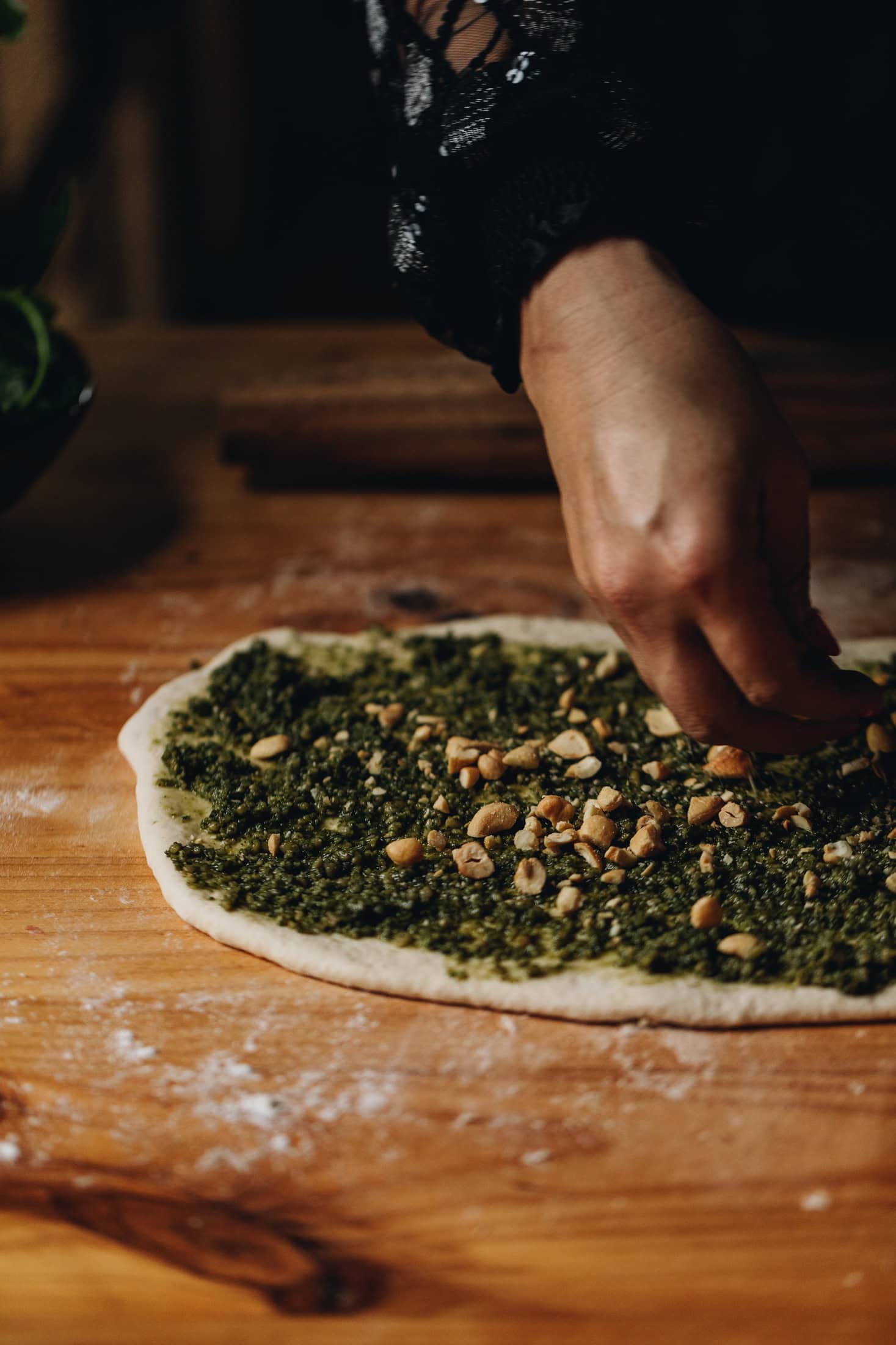 The rolled out cheese and pesto scrolls has been topped with pesto and is being sprinkled with chopped cashew nuts with a hand.