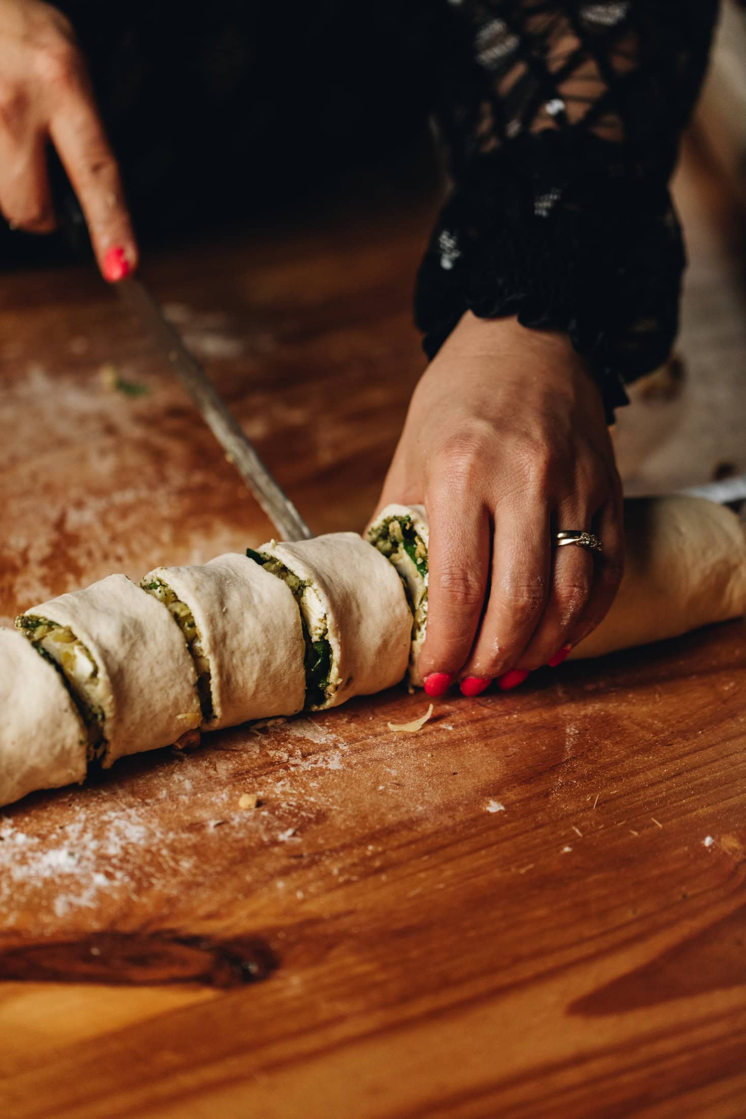 The cheese and tomato scroll dough is being cut in to 12 pieces on a wooden table with a knife
