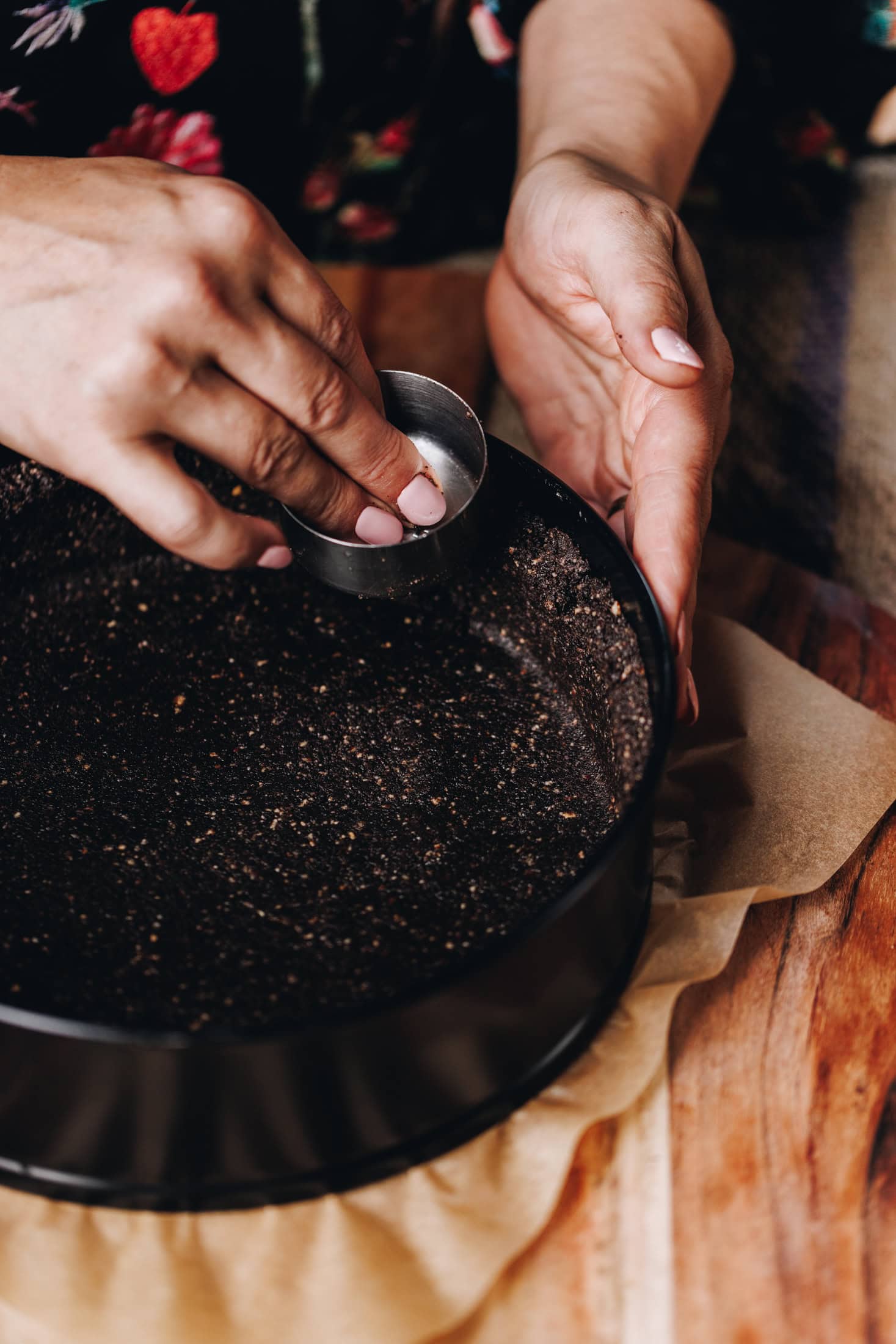 A black cake tin has the chocolate biscuit crumb in it. It is being pressed in to the tin by a small measuring cup. It is on a wooden table.