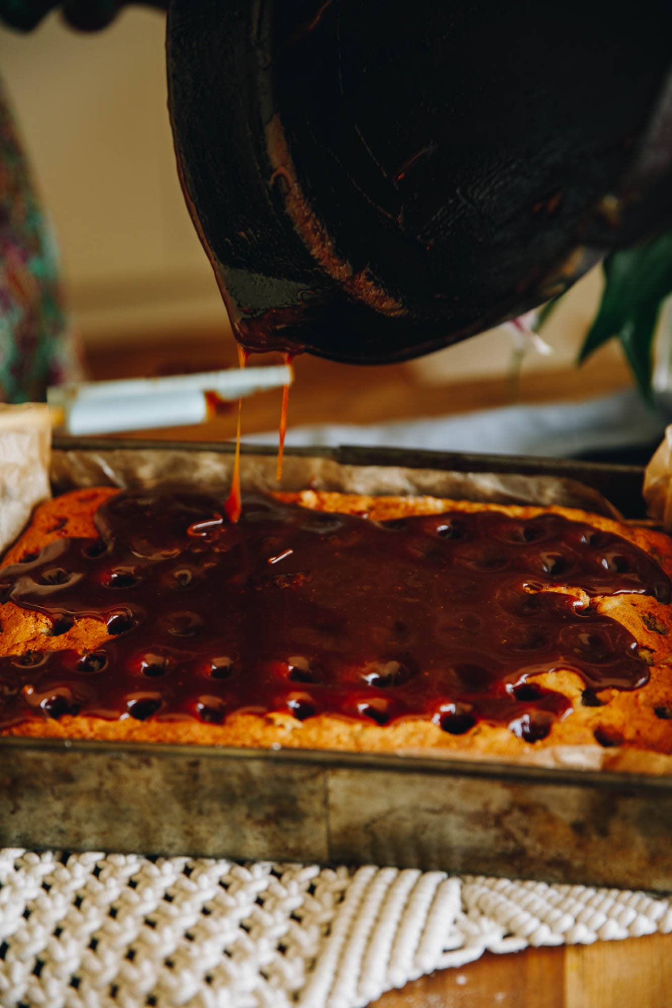 On a cream crocheted mat sits a freshly baked banana cake that has holes all over the top of it. Deep coloured caramel has been poured on top and the drips from a black cast iron pot are in view. 