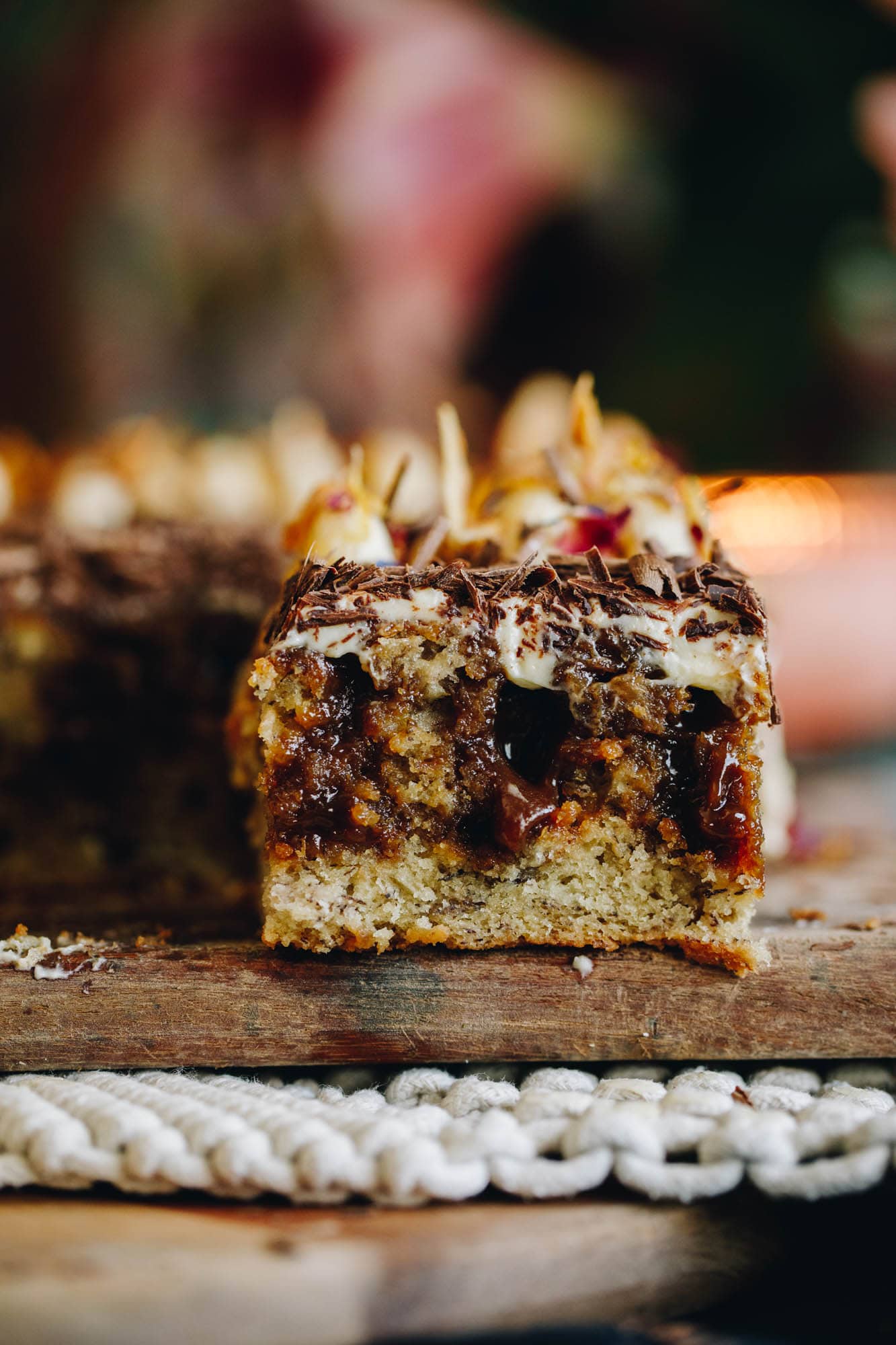On a wooden board sits a slice of banana and caramel poke cake that reveals the gooey caramel that has been poked inside the cake. On top is grated chocolate and cream cheese frosting with coconut chips. 