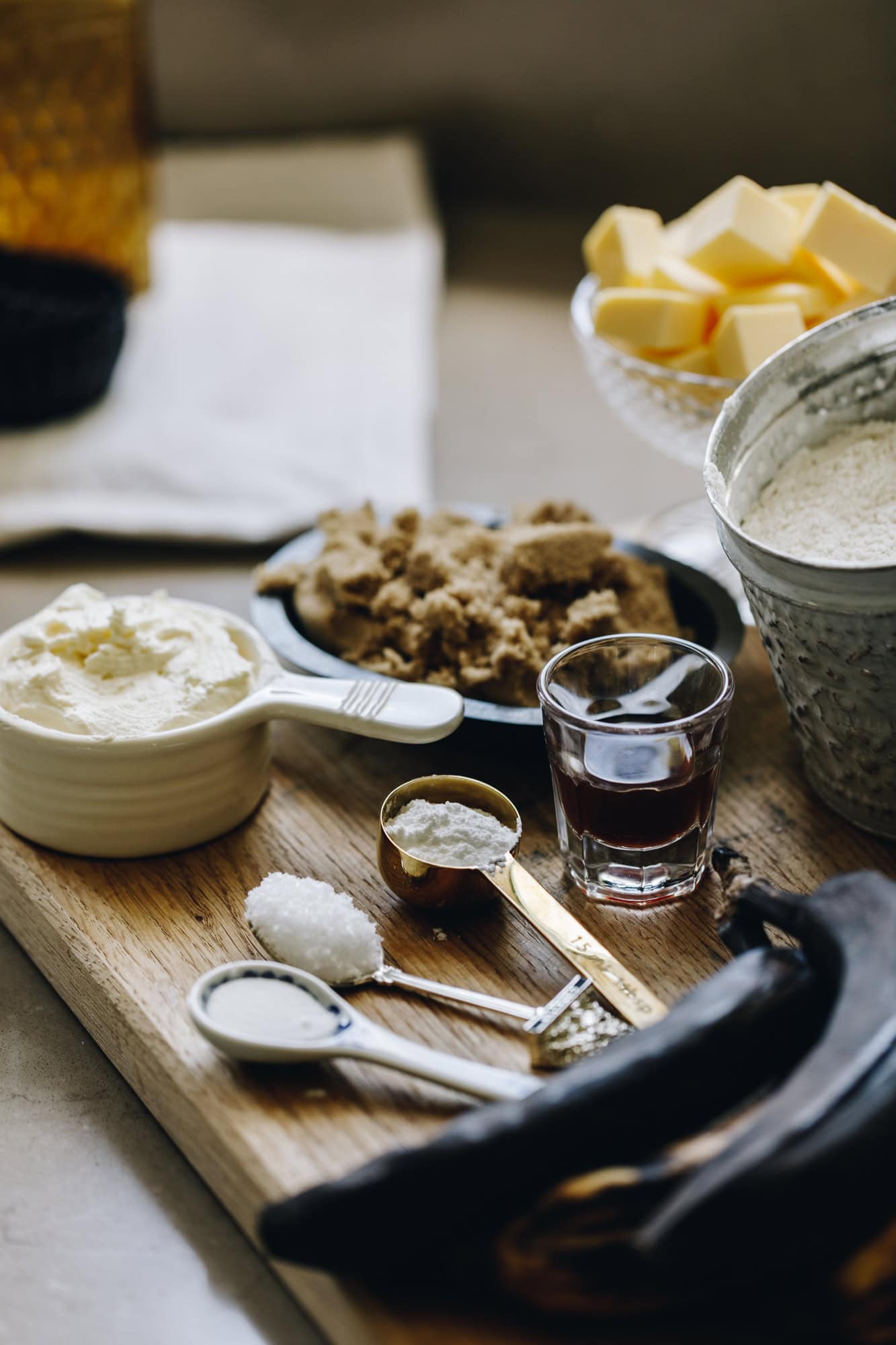Cake ingredients are in different vintage bowls and are sitting on a wooden board on top of stone bench. There is bananas, salt, yogurt, brown sugar and butter in shot.