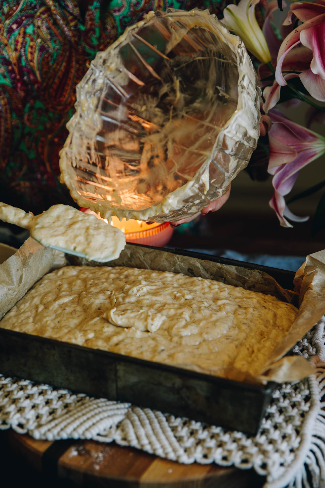 A baking tin lined with baking paper is sitting on a cream crocheted mat. A bowl has just finished pouring in the banana cake mixture with a blue spatula. 