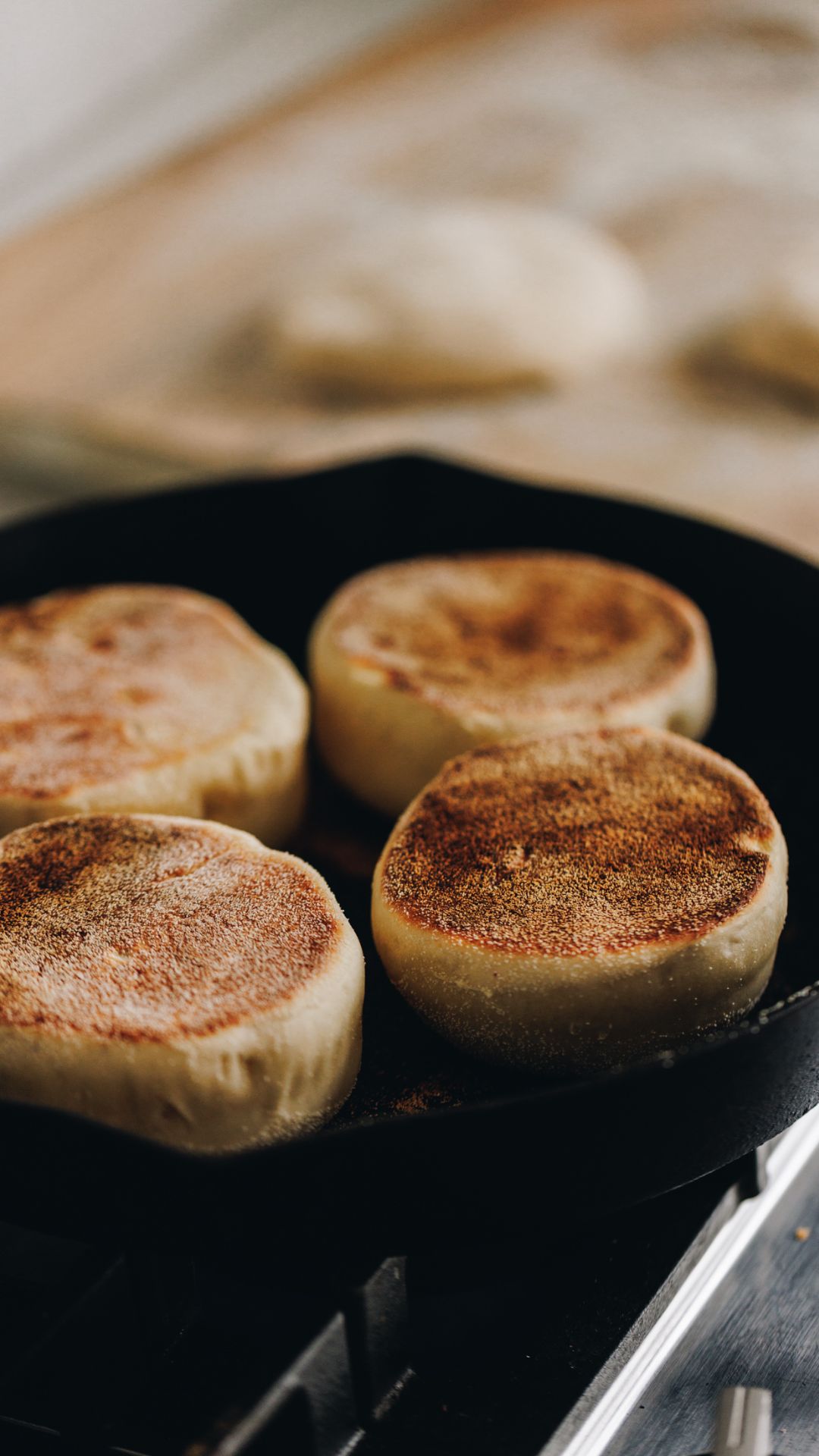 In a black cast iron pan is four homemade muffin splits cooking. The tops of a deep golden colour and in the background is a tray lined with brown baking paper and unbaked muffin split dough on it.