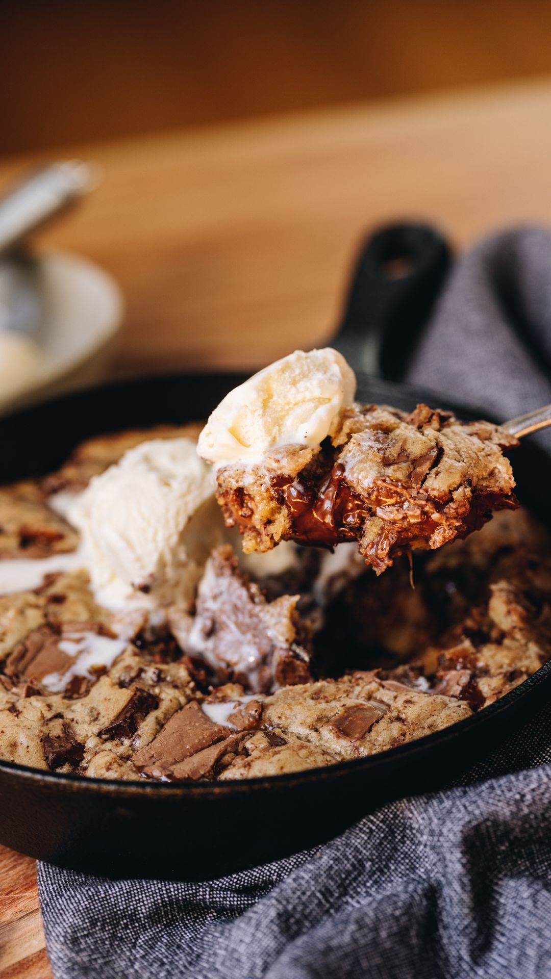 A black cast iron pan sits on top of a blue tea towel, on a wooden table. In the cut iron pan is a freshly baked chocolate caramel skillet cookie that has melty ice cream in the centre. Naomi is lifting some of the cookie with a silver spoon and it shows the gooey centre.