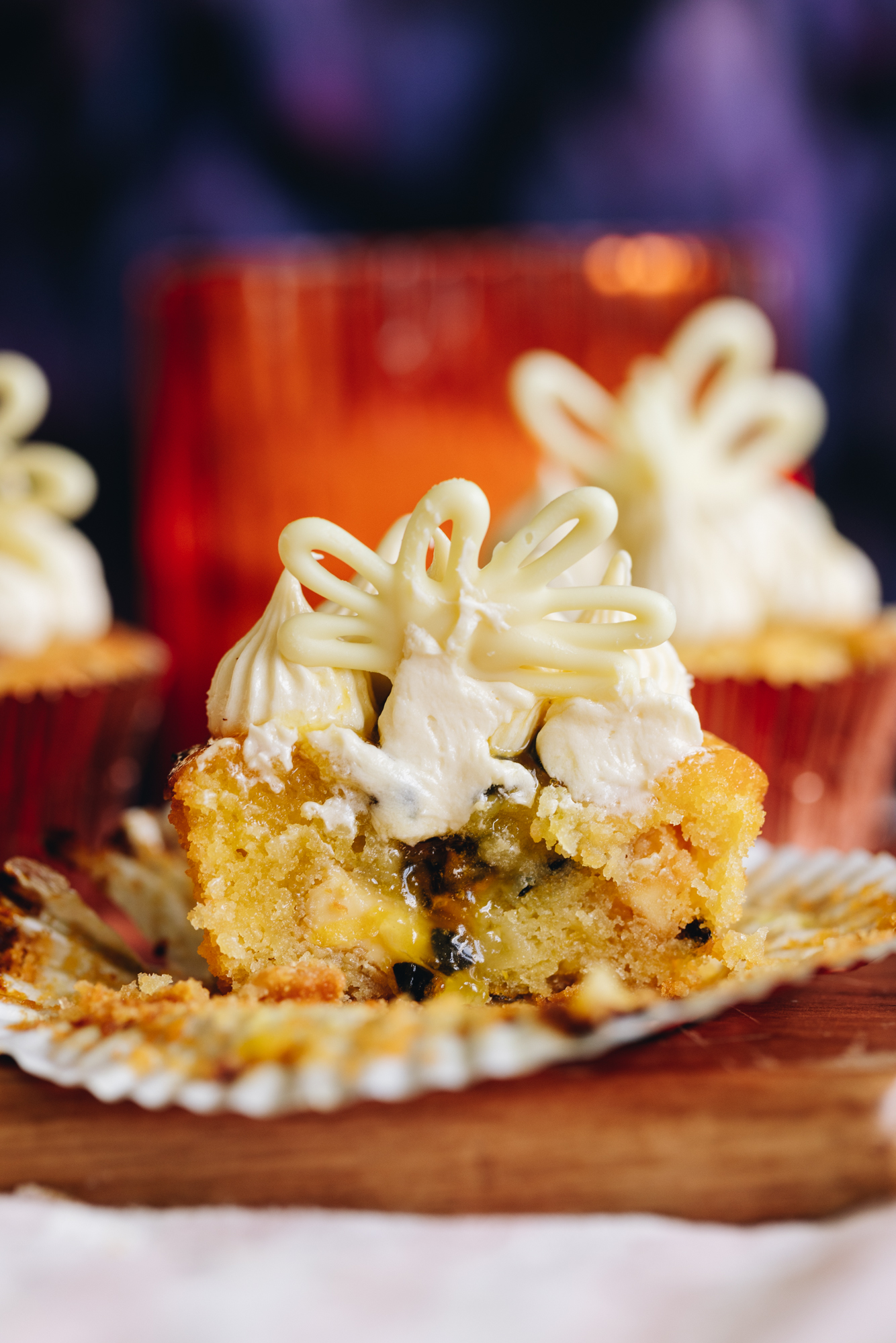A passionfruit white chocolate muffin is cut open to reveal the gooey passionfruit centre. It is topped with a cream cheese frosting and a decoration. There are other muffins in the background.
