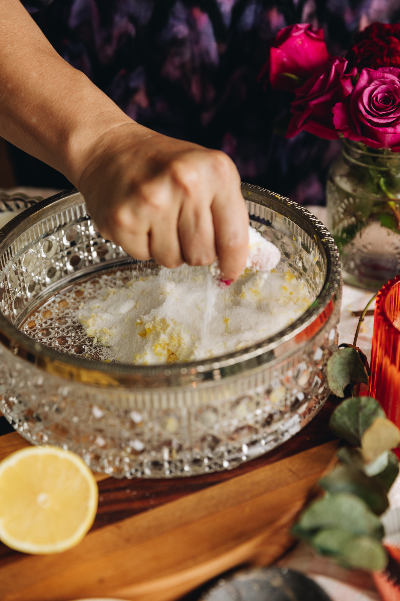 A glass vintage bowl is on a striped wooden board. In the bowl is caster sugar with lemon zest that is being mixed together by Naomi's hand. Purple flowers are in the background. 