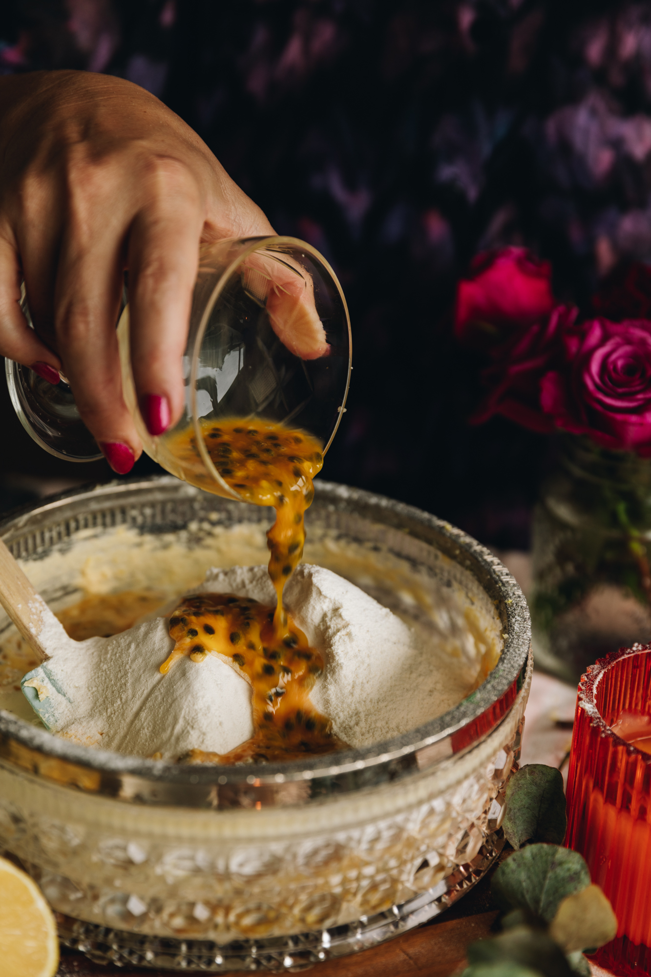 A glass vintage bowl is on a round wooden board. Whipped butter and sifted flour are in the bowl and Naomi is pouring passionfruit pulp in to the bowl. Purple flowers are in the background. 