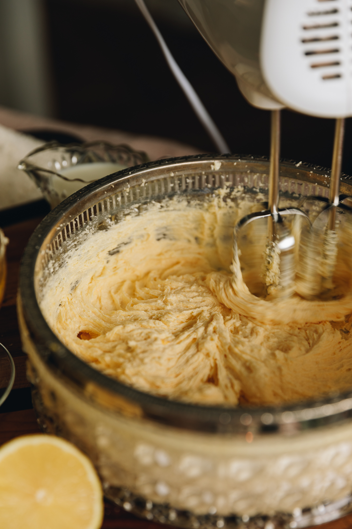 A glass vintage bowl is on a round wooden board on a grey tablecloth. Butter and sugar is being whipped with a white hand mixer.